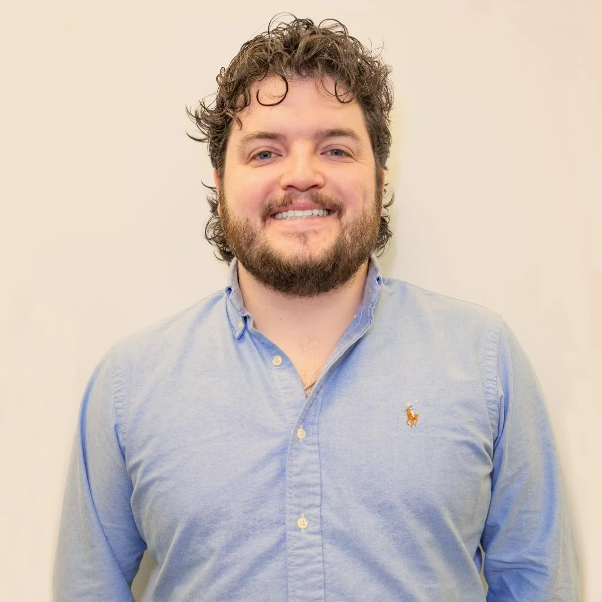 A smiling man with dark curly hair and a beard, wearing a light blue button-down shirt, standing against a plain beige wall.
