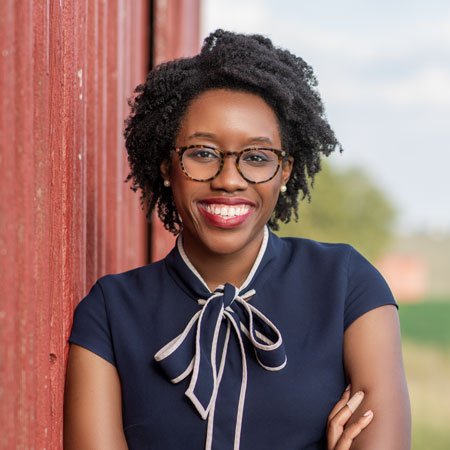 A smiling young woman with curly black hair, glasses, and a navy blue blouse with a bow, standing outdoors beside a red wooden wall.