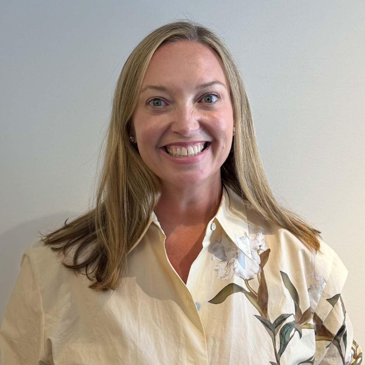 A woman with long blonde hair smiling, wearing a cream-colored floral blouse, standing against a plain light-colored wall.