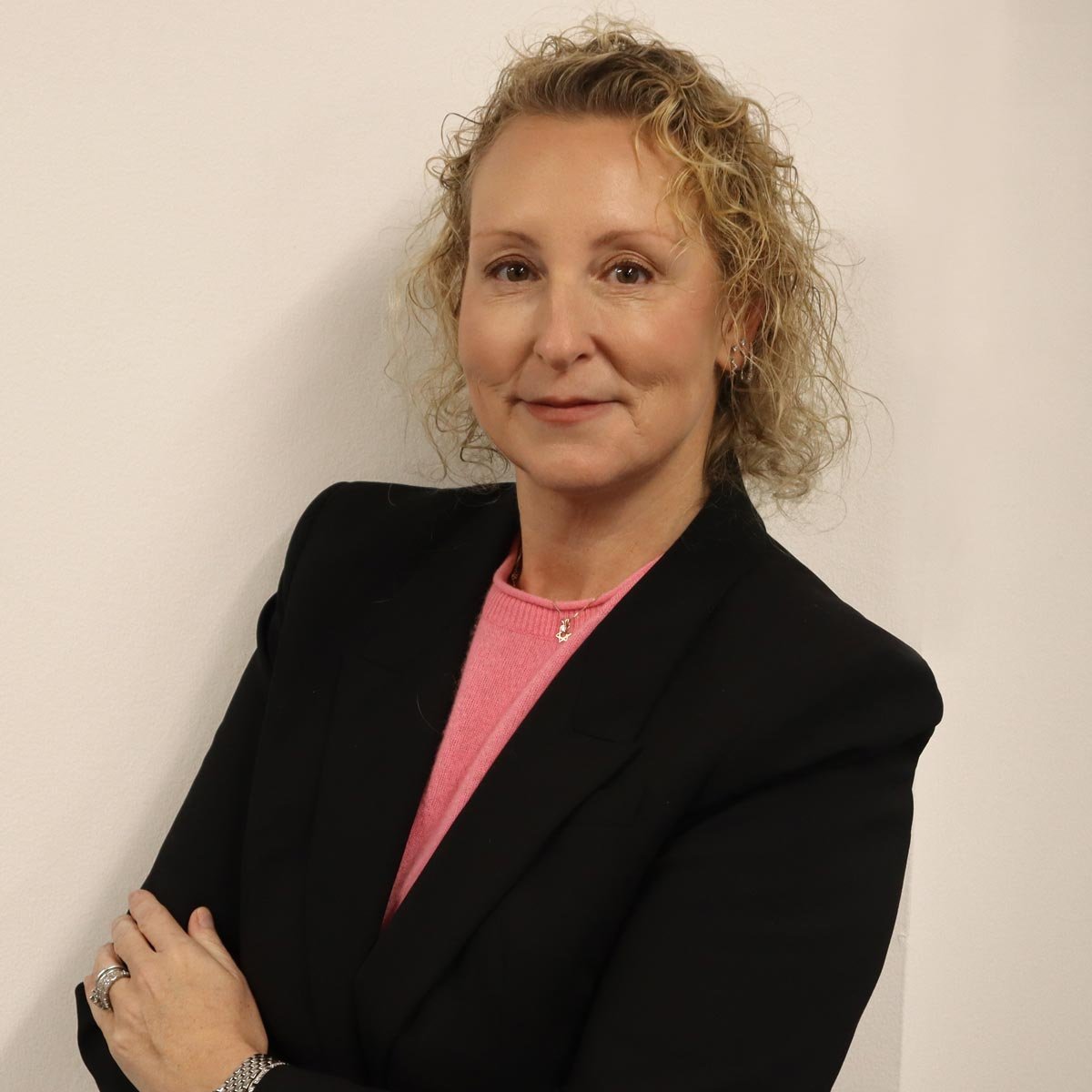 A woman with curly blonde hair wearing a black blazer and pink top, standing with her arms crossed against a plain white wall.