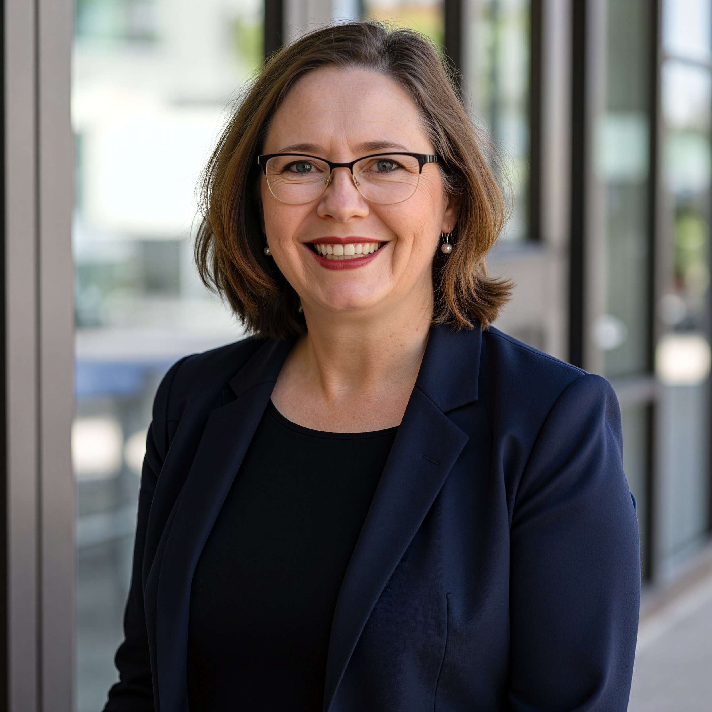 Portrait of a woman with shoulder-length brown hair, wearing glasses, earrings, a navy blazer, and a black top, standing in front of a glass door or window.