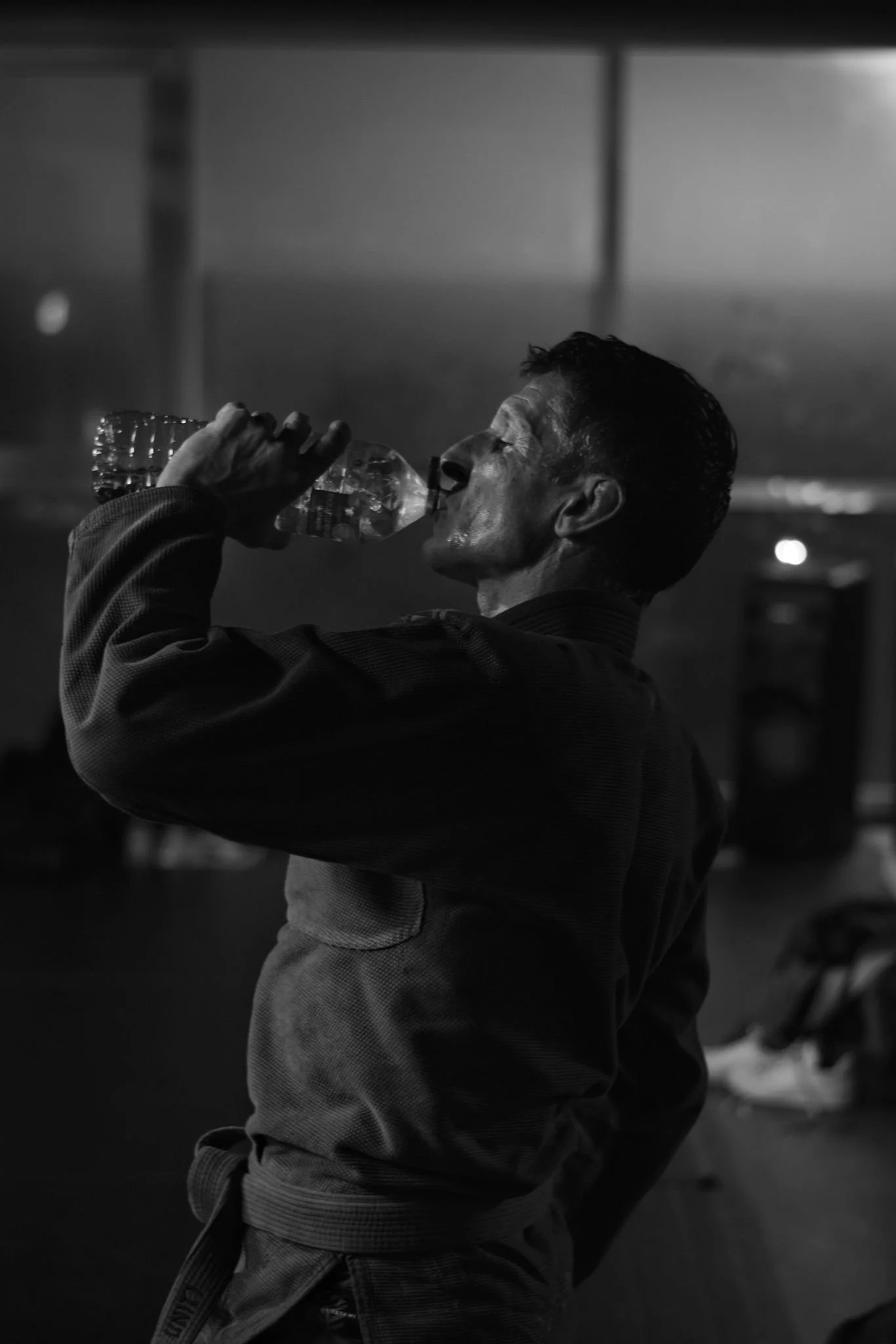 Un homme en kimono de jiu jitsu bleu clair boit de l'eau d'une bouteille dans une pièce sombre