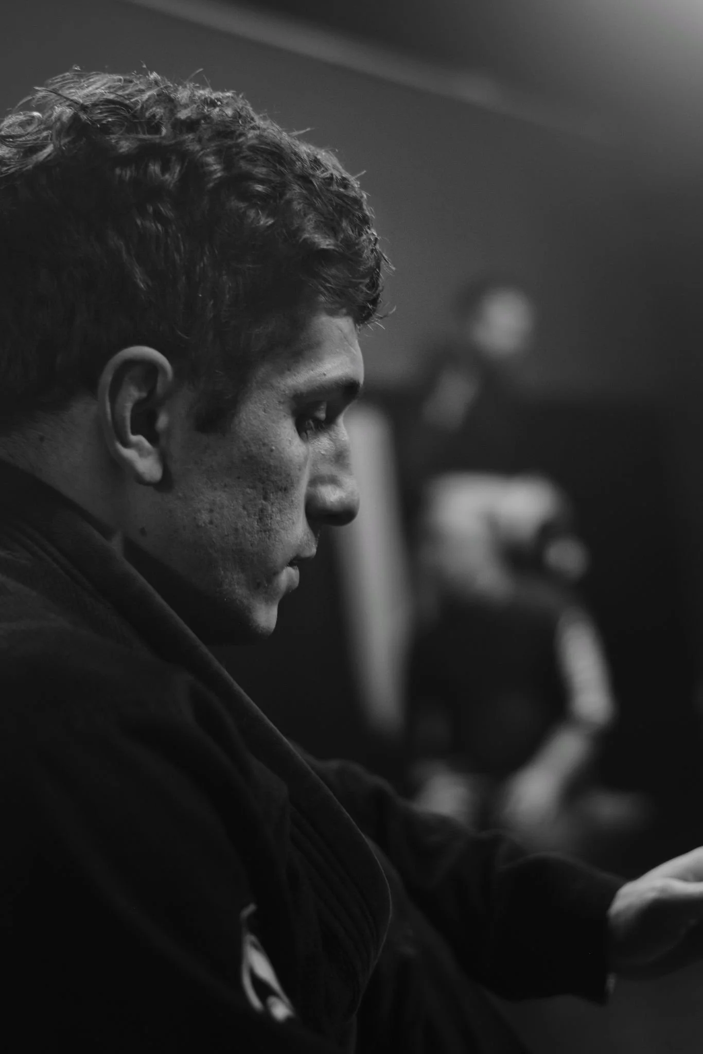 Un jeune homme en profil, concentré, en noir et blanc, portant un kimono de jiu jitsu, avec une autre personne floue en arrière-plan.