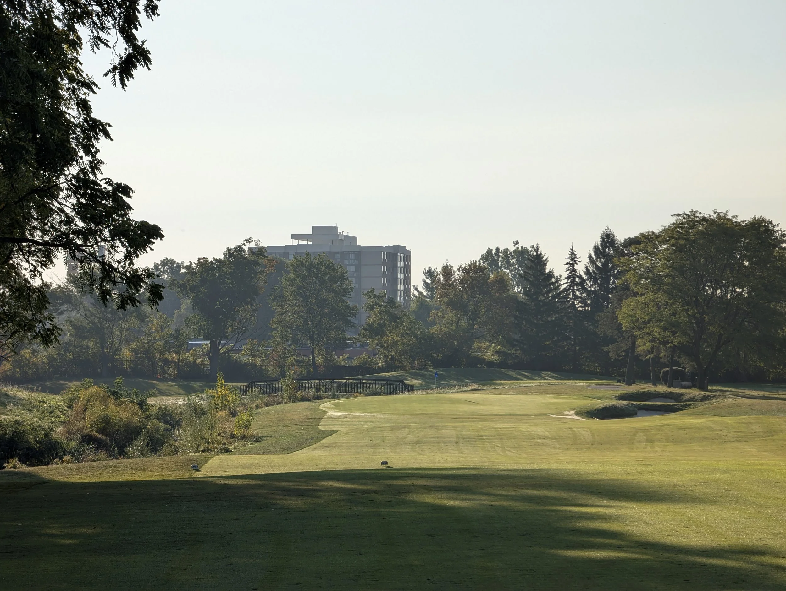 View of a golf course with a fairway and sand traps, trees lining the course, high-rise buildings in the background, and a clear sky.