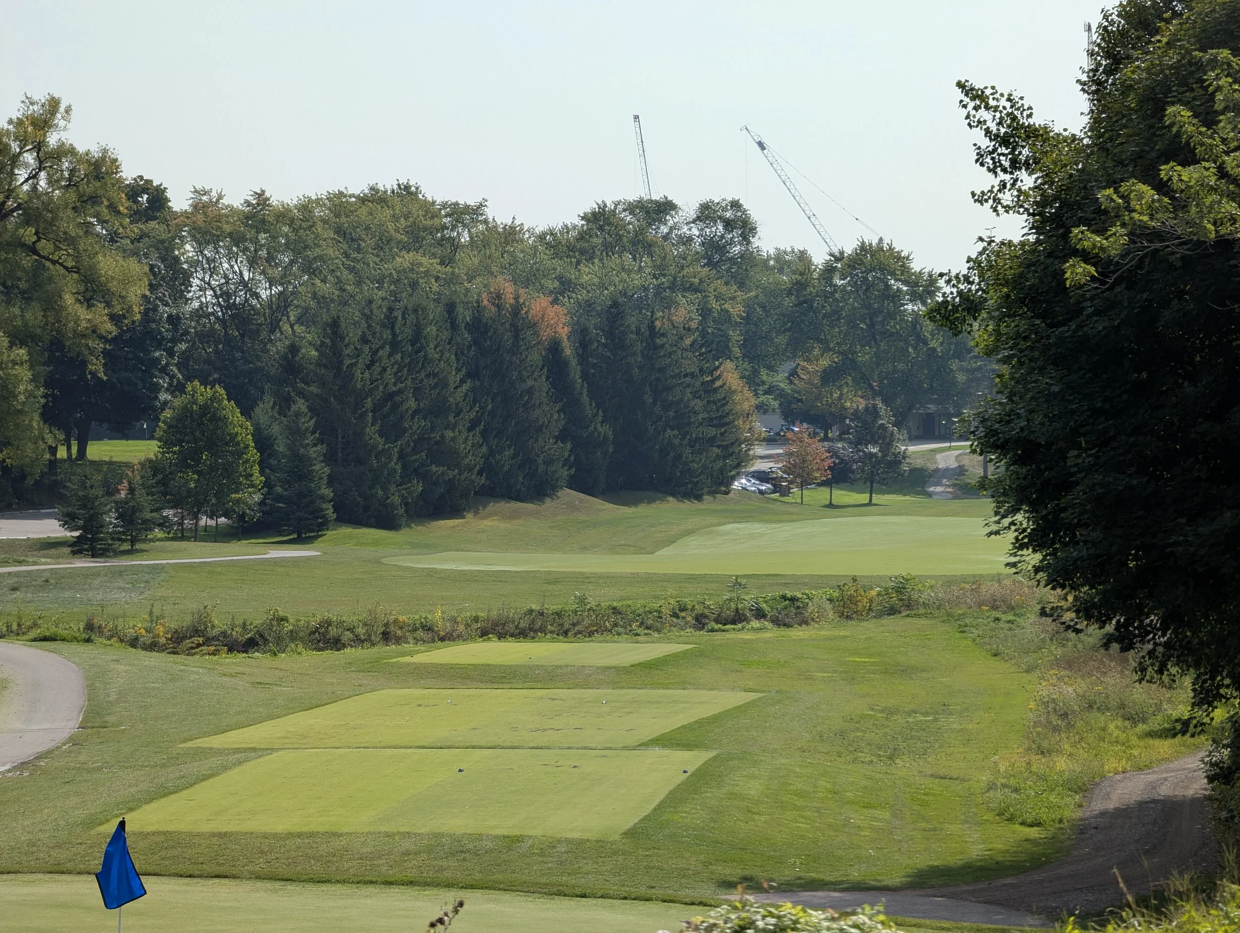 A golf course with a blue flag, well-maintained grass, trees in the background, and a construction crane in the distance.