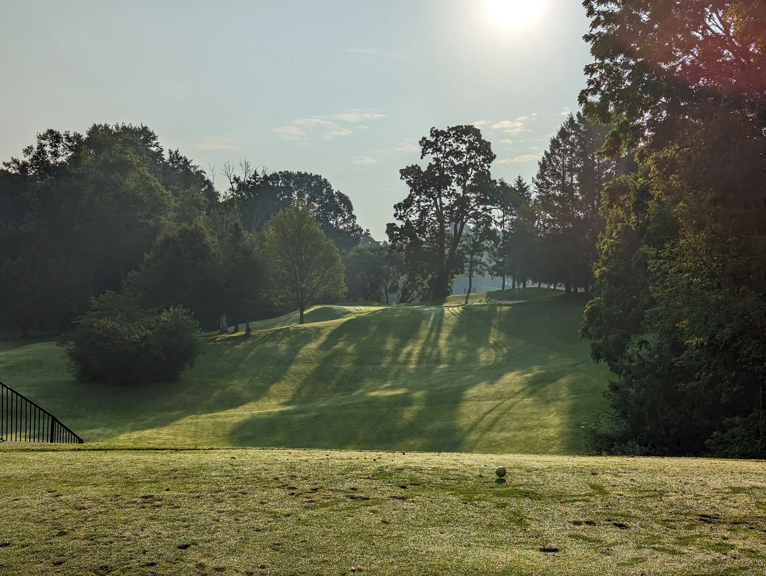 A golf course with a tee and a golf ball in the foreground, grassy hills, trees, and sunlight creating shadows in the background.