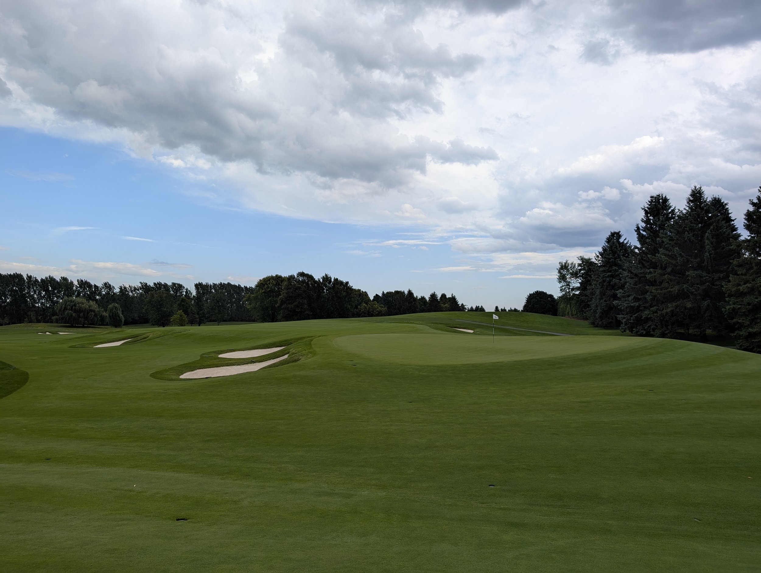 A lush green golf course with sand bunkers and a flagstick on the putting green, surrounded by trees and under a cloudy sky.