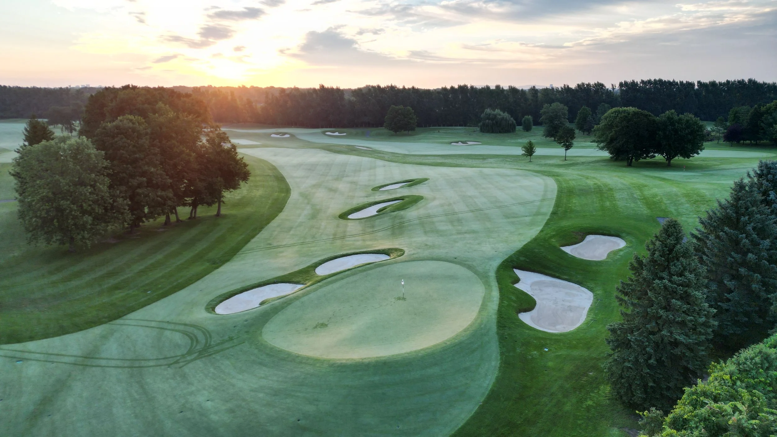 Aerial view of a golf course during sunset, showcasing green fairways, sand bunkers, and trees.