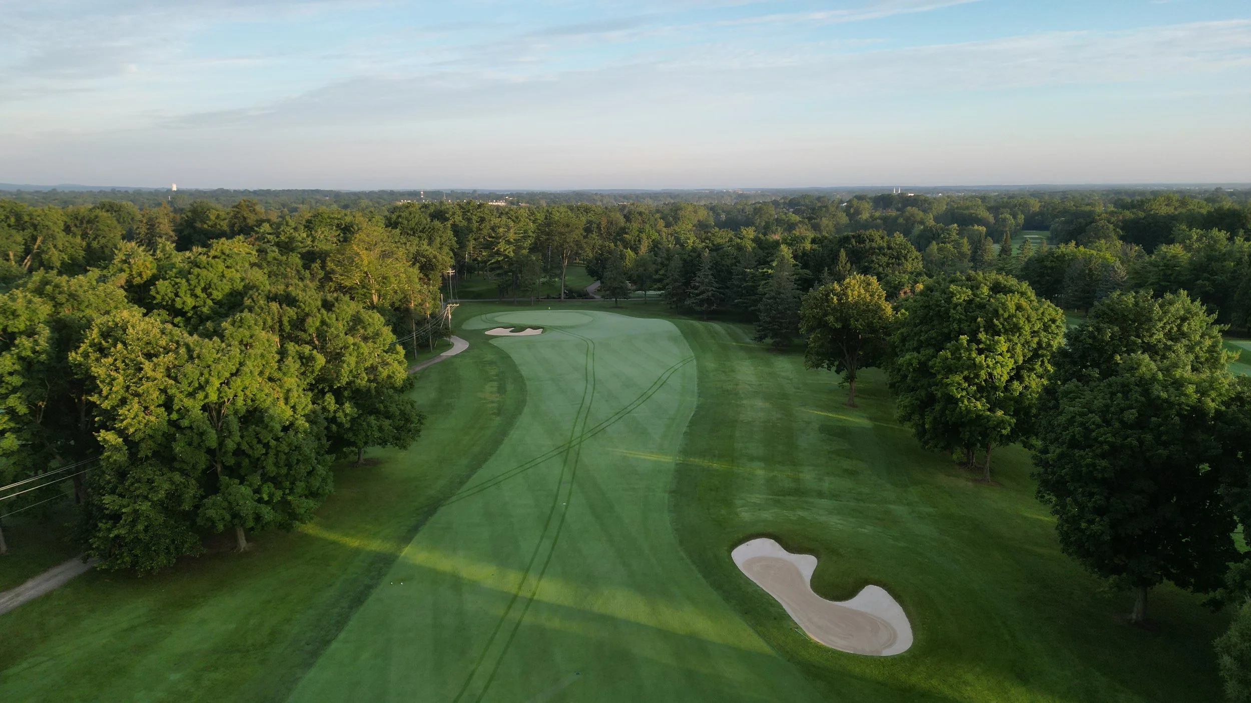 Aerial view of a golf course with green fairway, sand bunkers, and trees on both sides, under a partly cloudy sky.