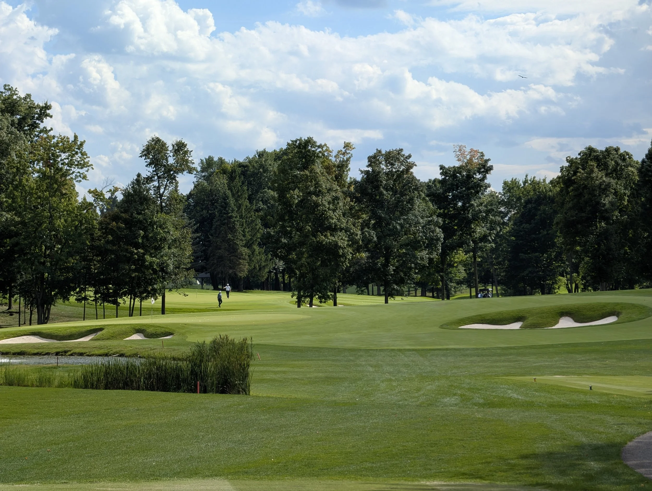 A golf course with well-maintained grass, sand bunkers, and trees in the background under a partly cloudy sky.
