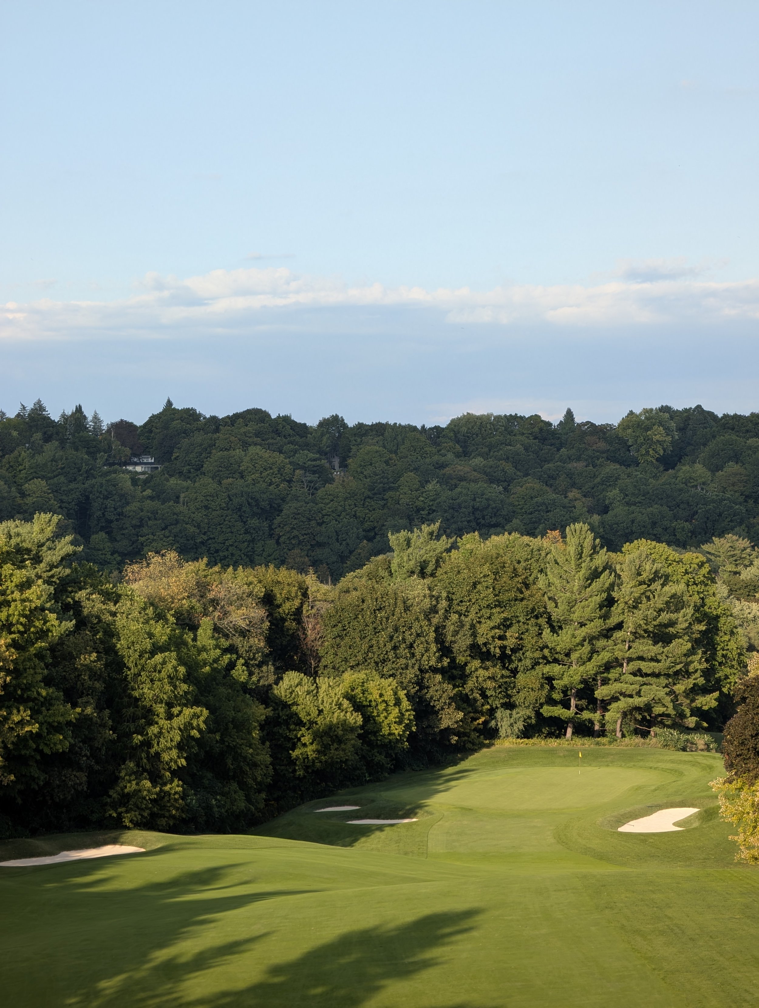 A golf course with a green, sand bunkers, surrounded by trees, under a partly cloudy sky.