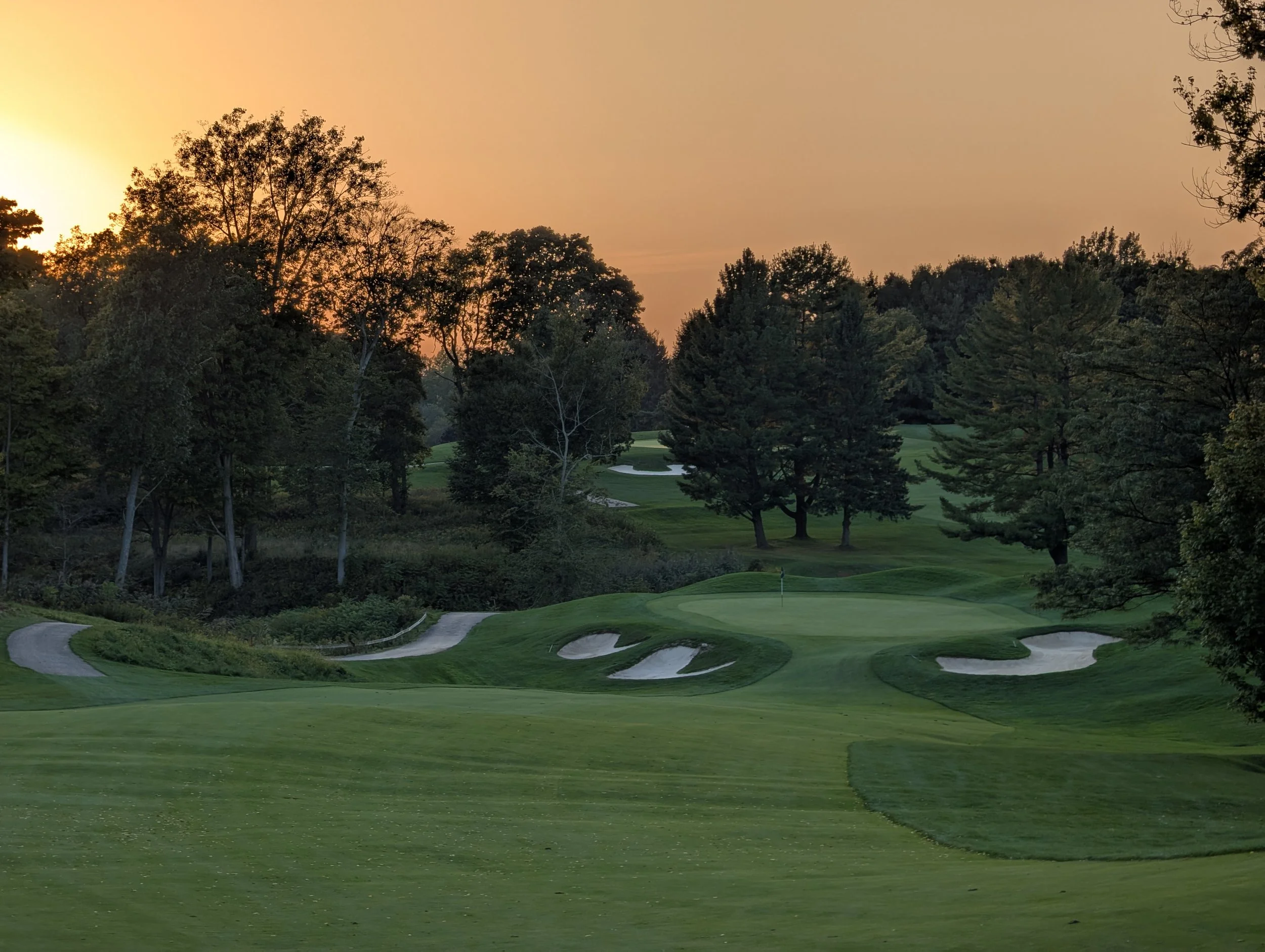 A golf course at sunset with sand bunkers, lush green fairways, and trees surrounding the area.