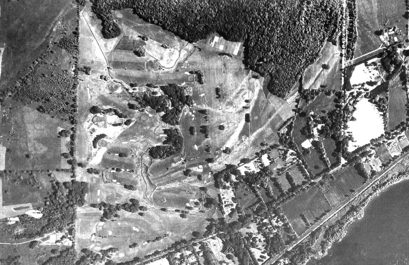 Aerial black and white photo of a rural landscape with fields, trees, winding roads, and scattered houses.