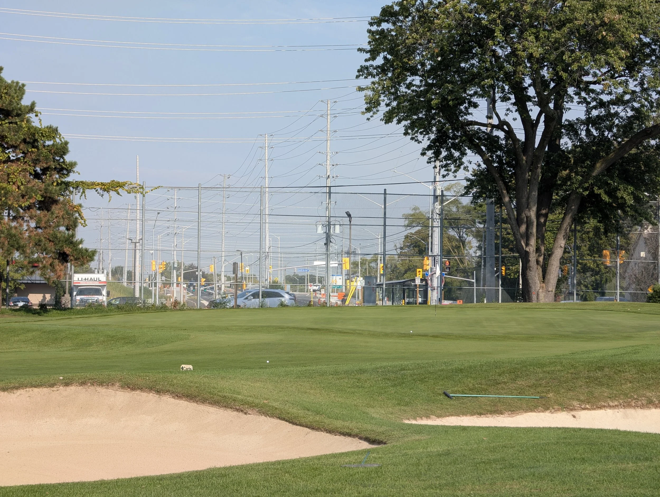 A golf course with green grass, sand bunkers, and a golf tee marker, surrounded by trees and a chain-link fence with a busy street and traffic in the background.