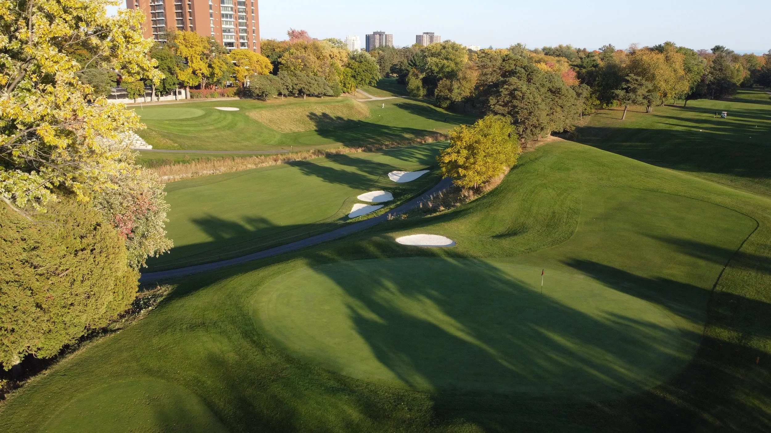 Aerial view of a golf course with green fairways, sand bunkers, and trees, with some buildings visible in the background.