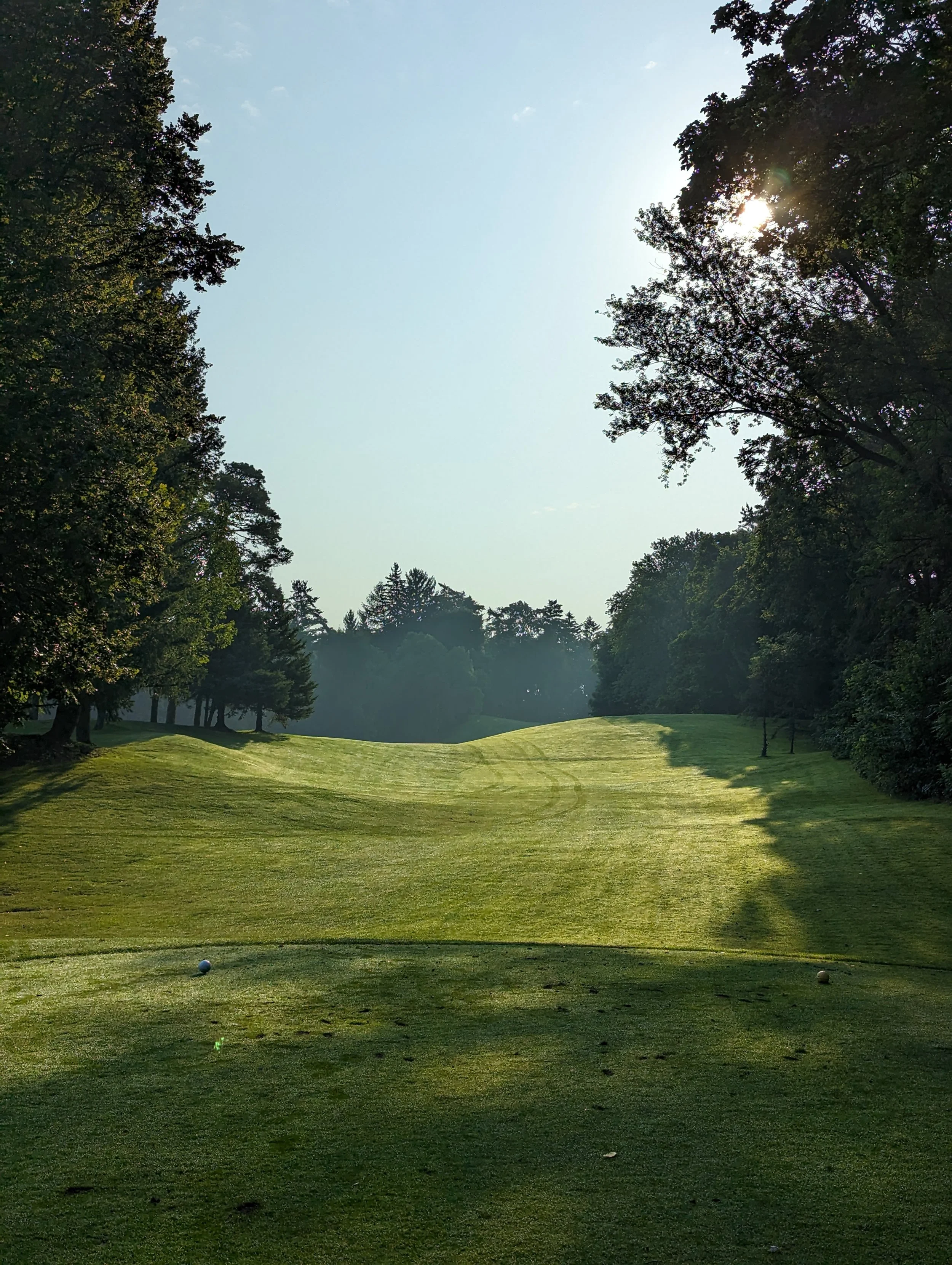 A golf course fairway with two golf balls, surrounded by trees under an early morning or late afternoon sky with the sun partially obscured by trees.
