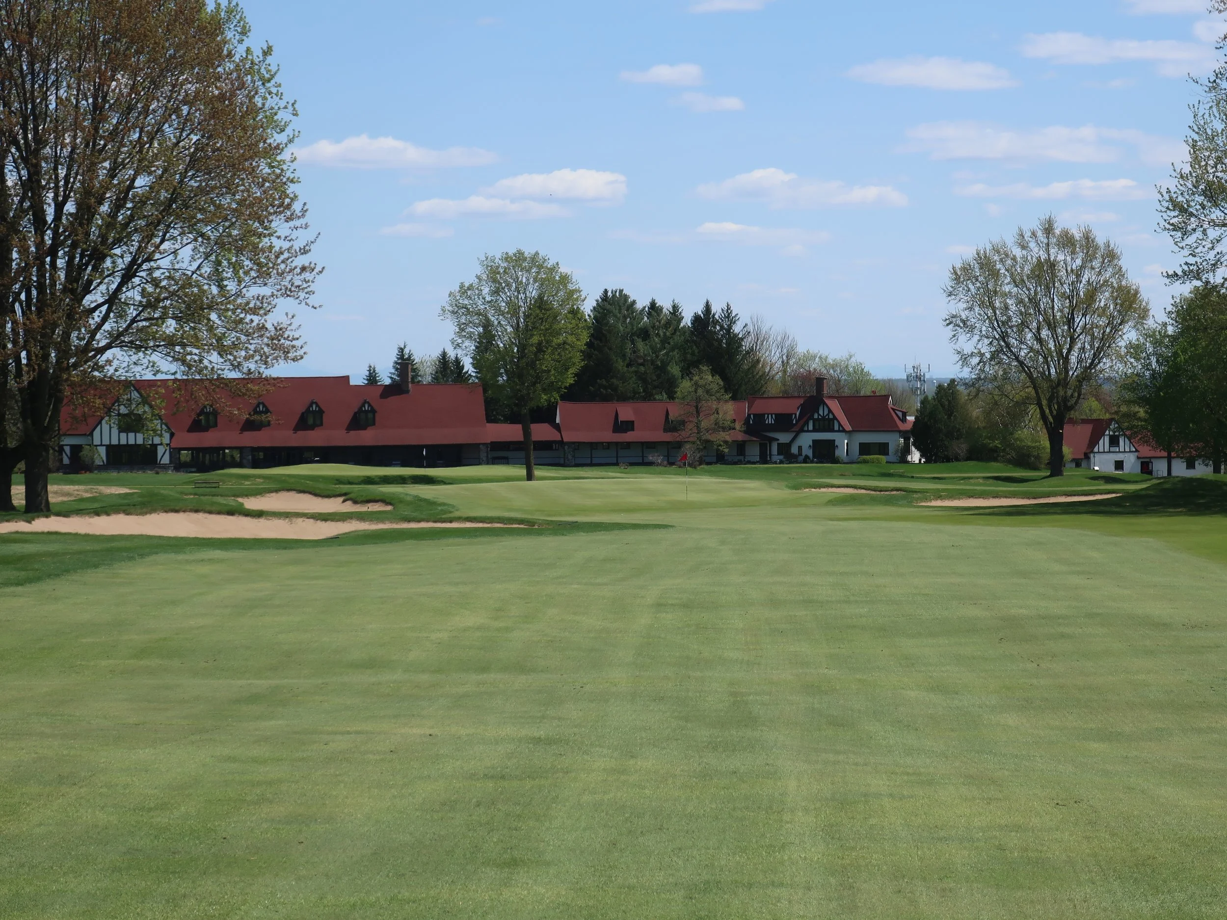 Golf course with a putting green, sand bunkers, and trees, with buildings with red roofs in the background under a partly cloudy sky.