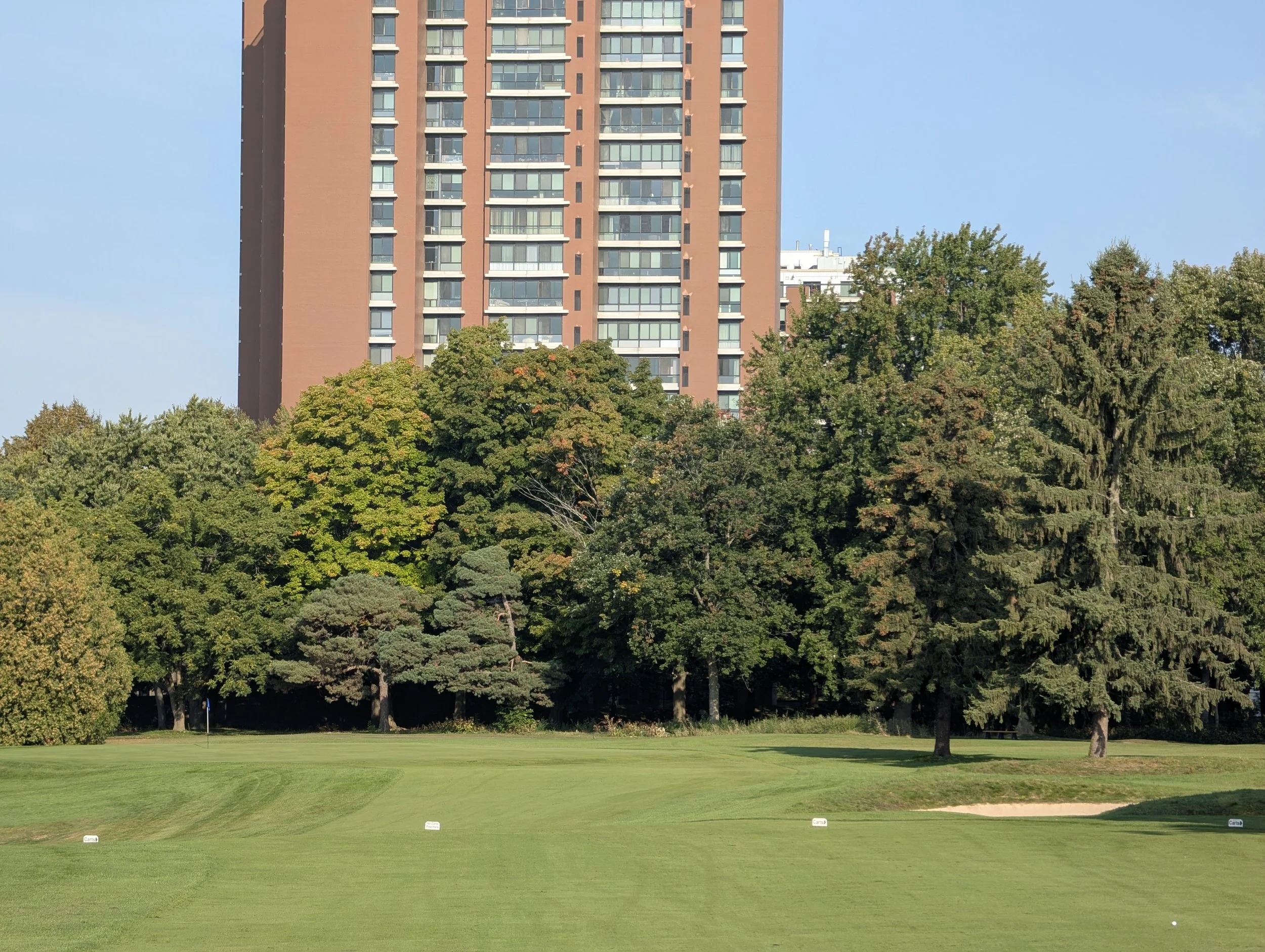 A golf course with green grass and a small sand trap, surrounded by trees and a tall residential apartment building in the background under a clear blue sky.