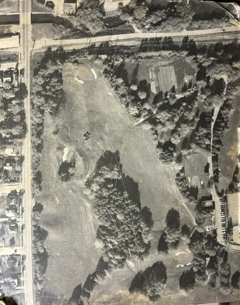 Aerial view of a golf course with fairways, greens, sand bunkers, trees, and surrounding roads and structures.