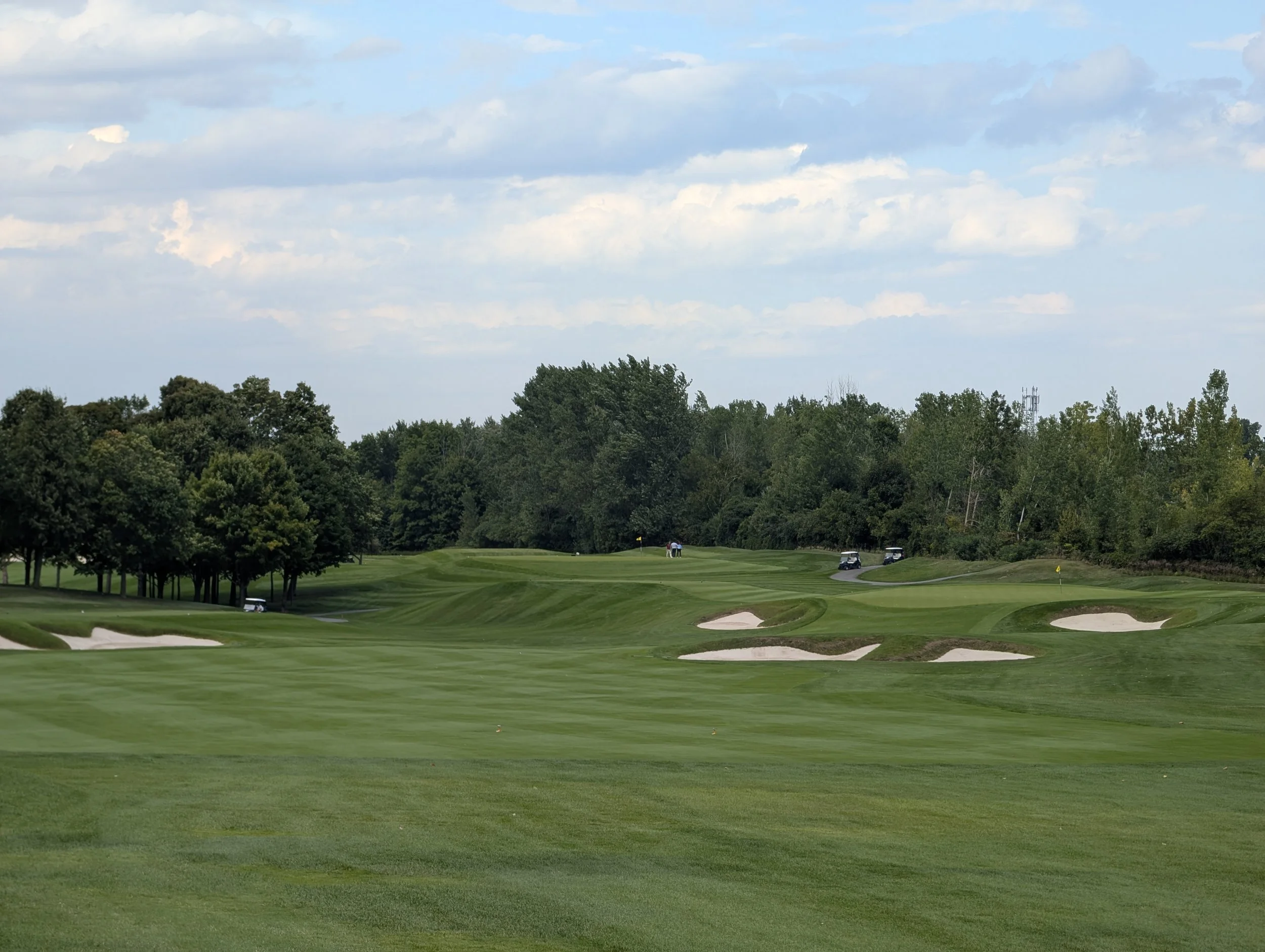 A golf course with well-maintained grass, sand bunkers, and trees on the perimeter under a partly cloudy sky.