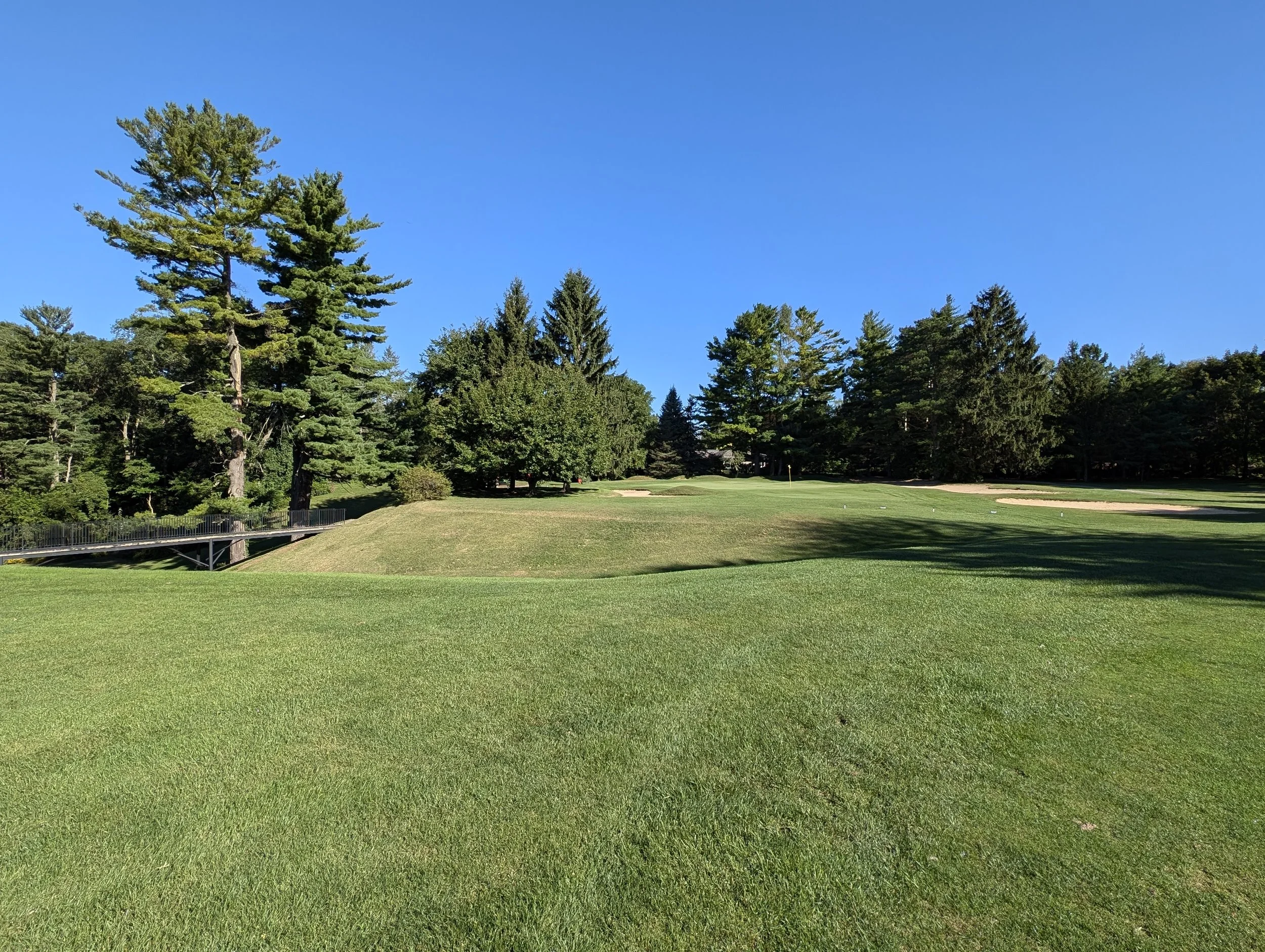 A golf course with well-maintained green grass, trees, and sand bunkers under a clear blue sky.