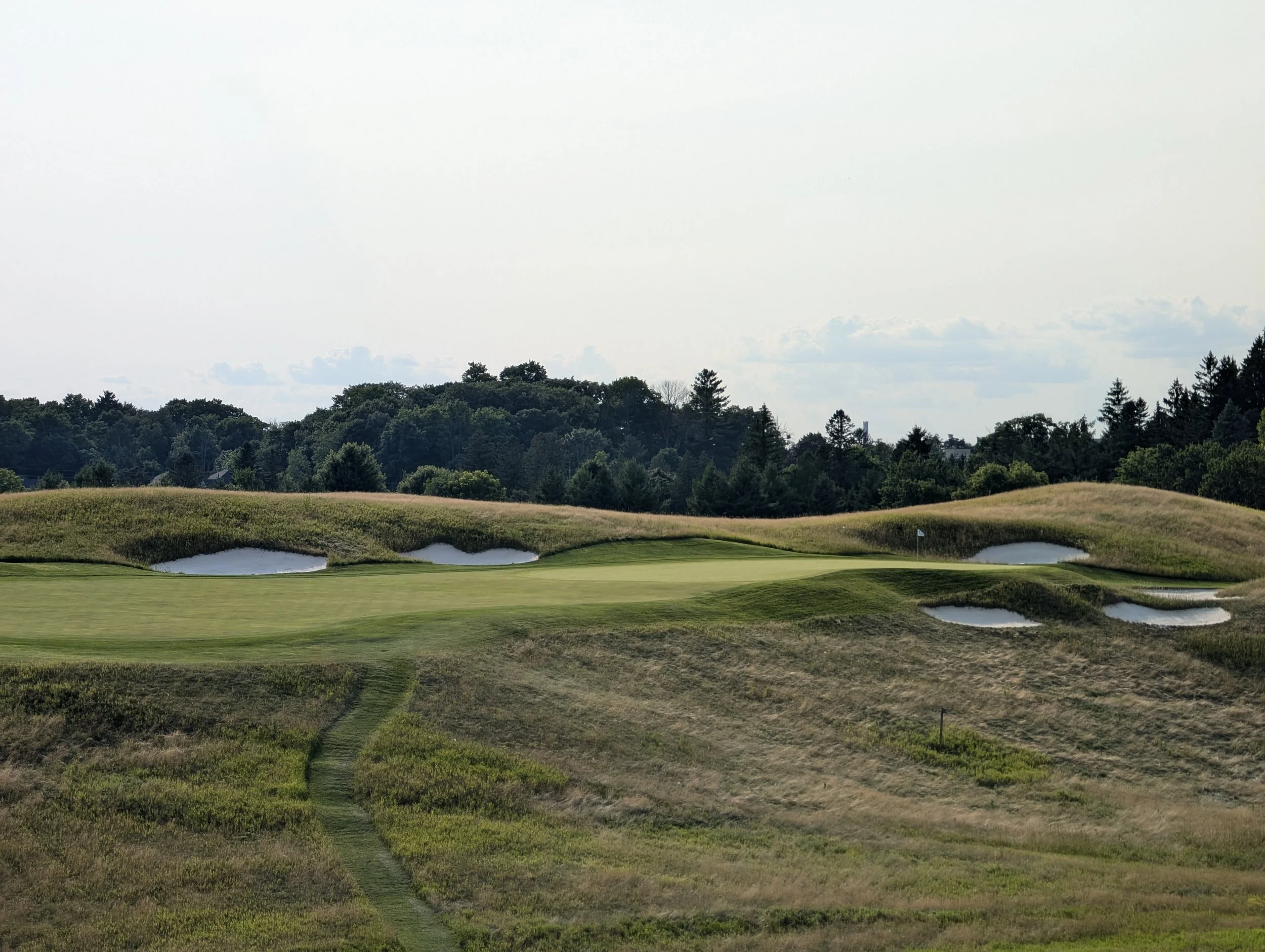 A golf course with green grass, sand bunkers, and a small body of water, surrounded by rolling hills and trees under a cloudy sky.