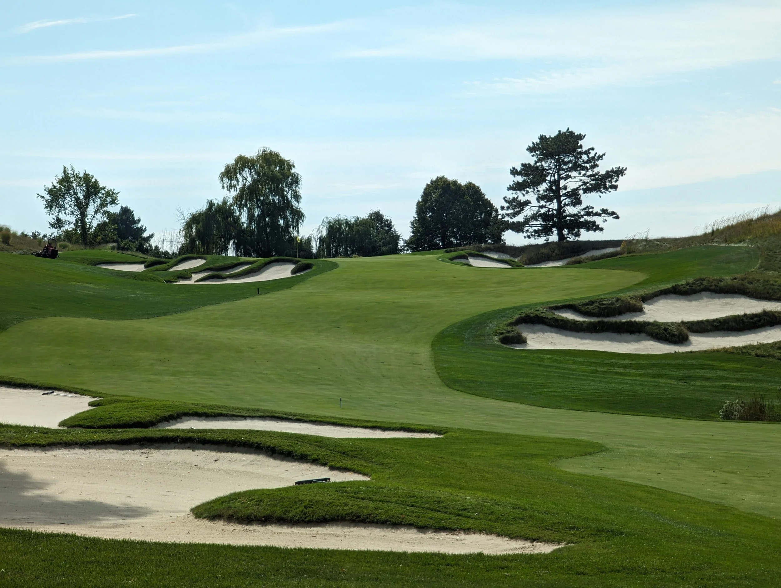 A scenic golf course with sand bunkers, lush green fairways, and trees under a partly cloudy sky.