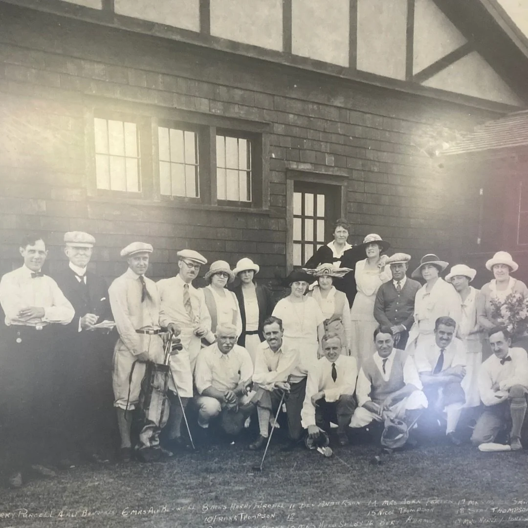 A black and white photograph of a group of men and women outdoors, standing and sitting in front of a building with wooden siding. Some are holding golf clubs, suggesting they are a golf team or club.