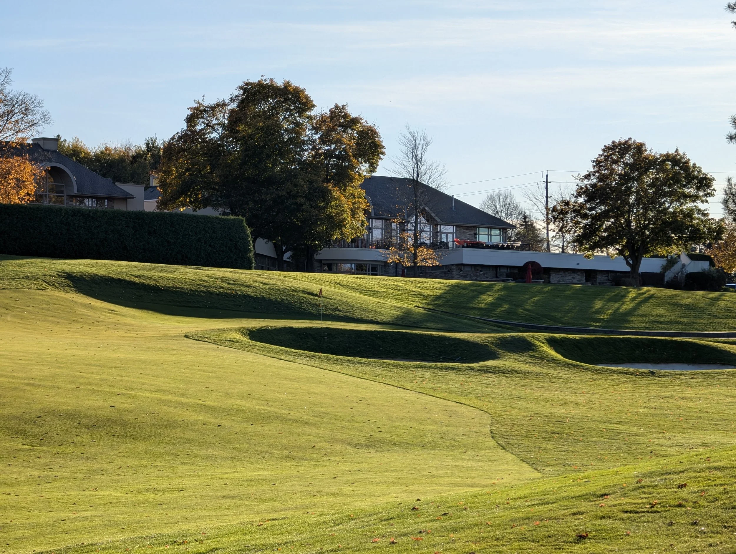 A golf course with well-maintained grass, small sand traps, and a flag marking the hole. In the background, trees with autumn leaves and residential buildings are visible under a clear sky.