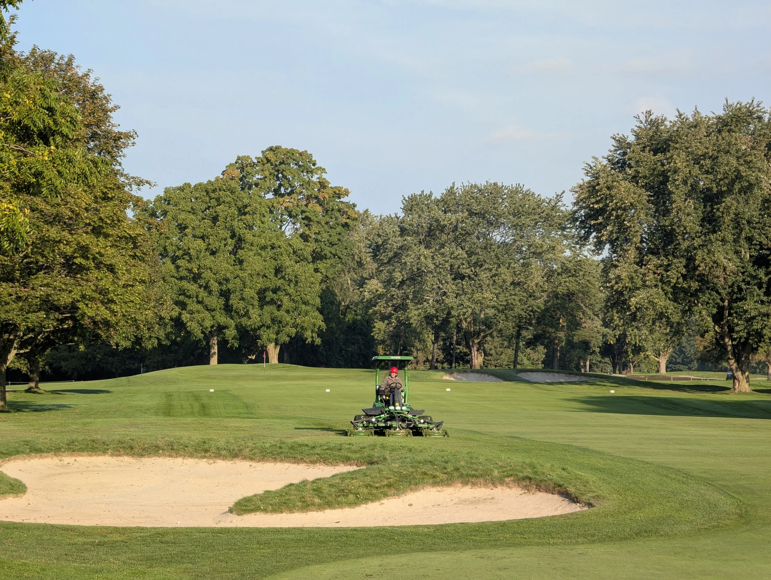 A person operating a golf course mower on a lush green fairway surrounded by trees.