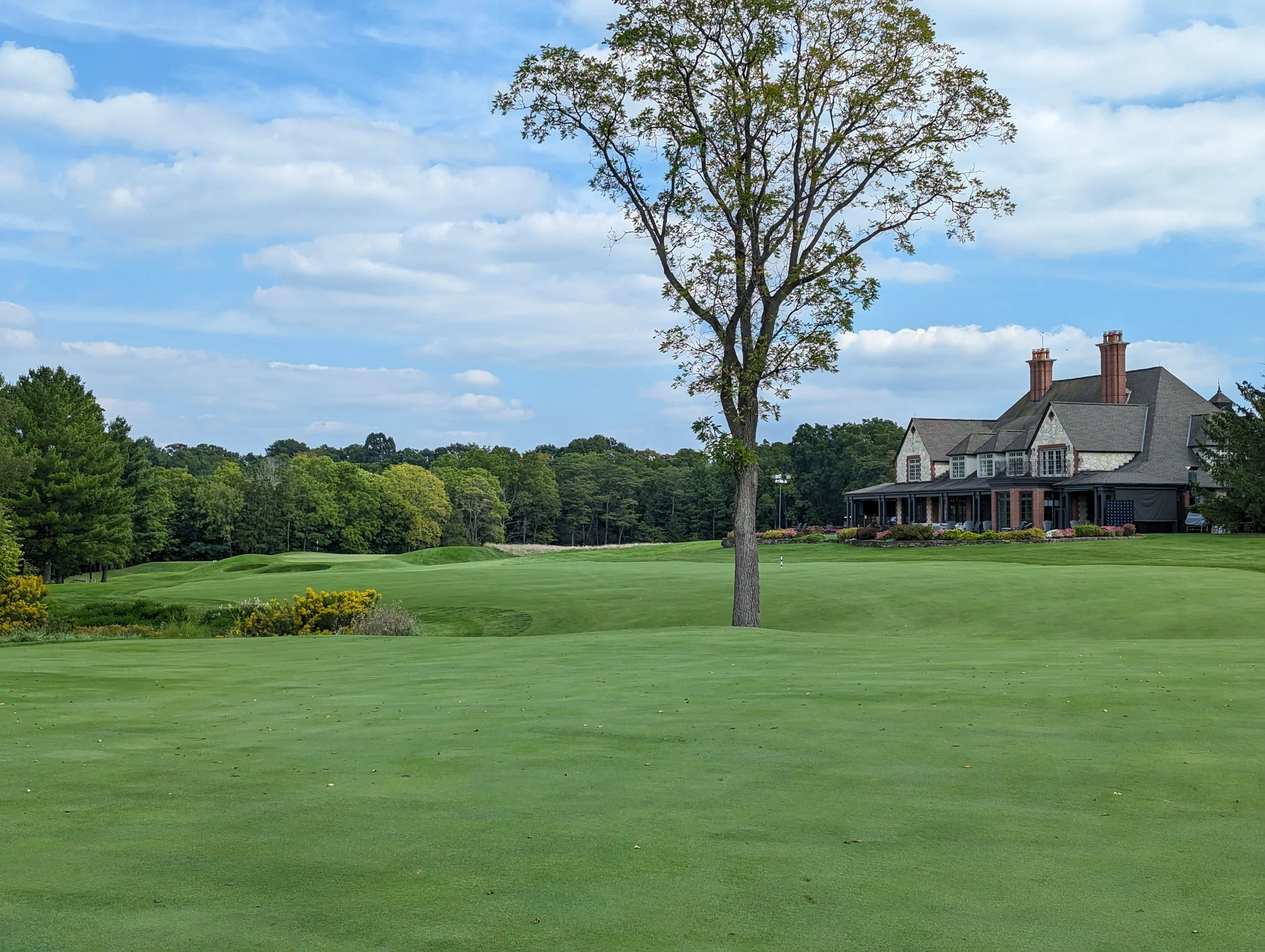 A lush green golf course with a large house in the background, tall trees, and partly cloudy sky.