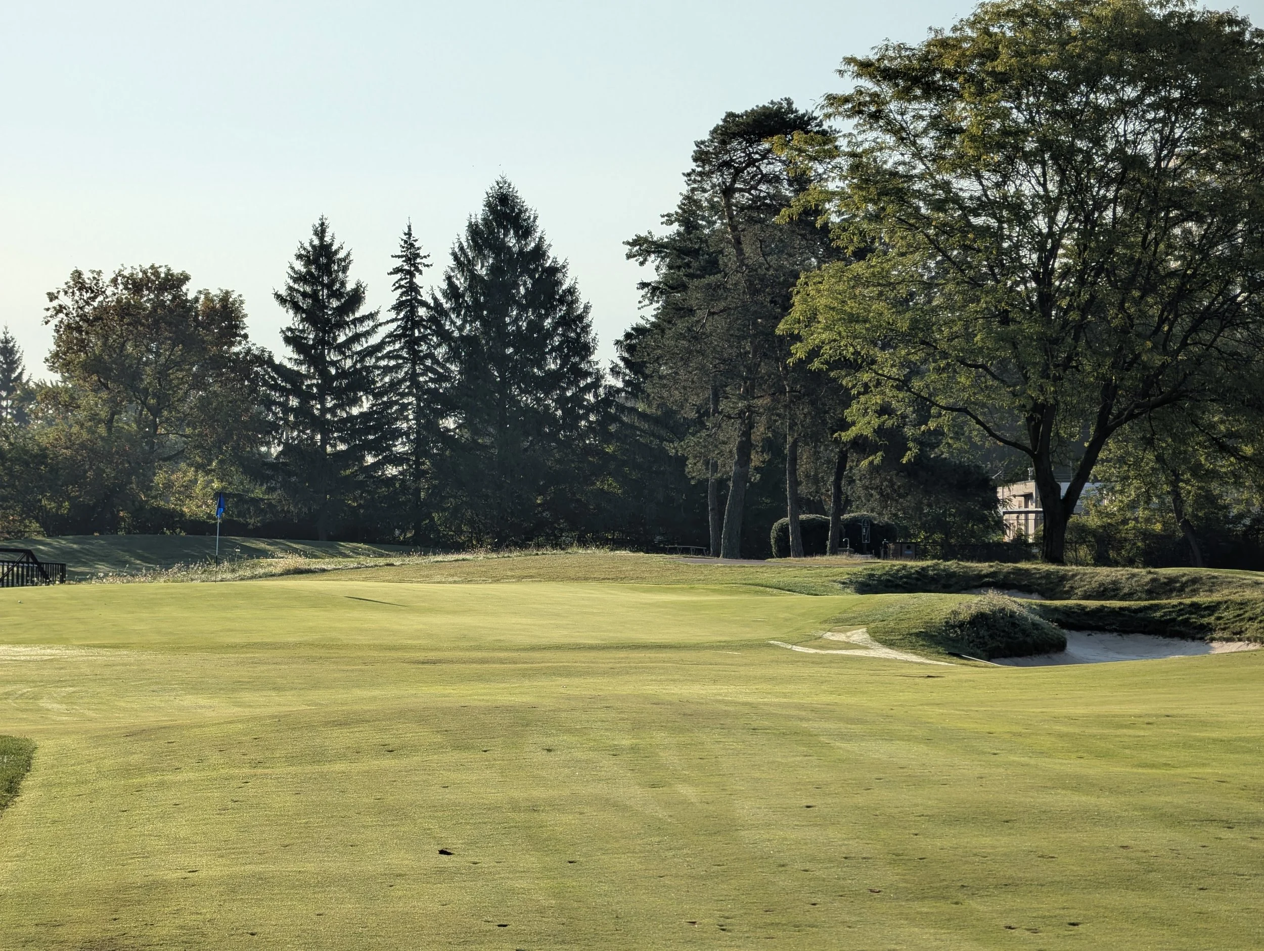 A golf course with green grass, a flag on a pole, and trees in the background on a sunny day.