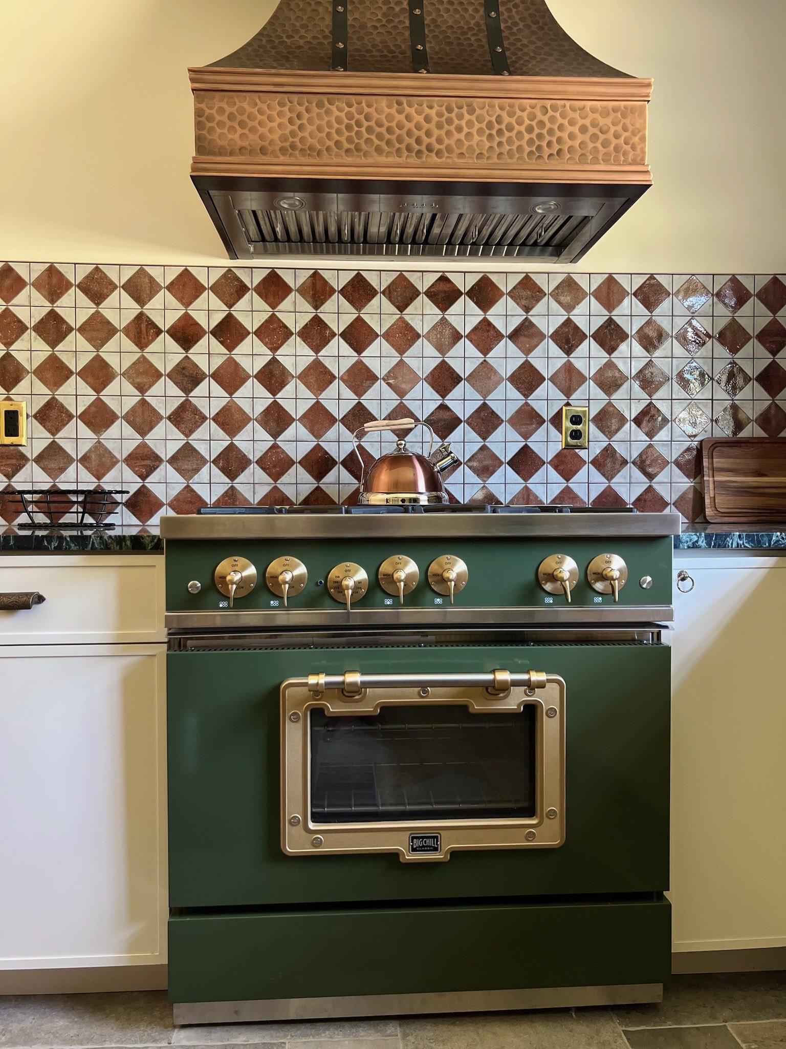 A green vintage-style kitchen stove with dials, a built-in oven, and a copper kettle on the stovetop, set against a patterned tile backsplash and a copper range hood.