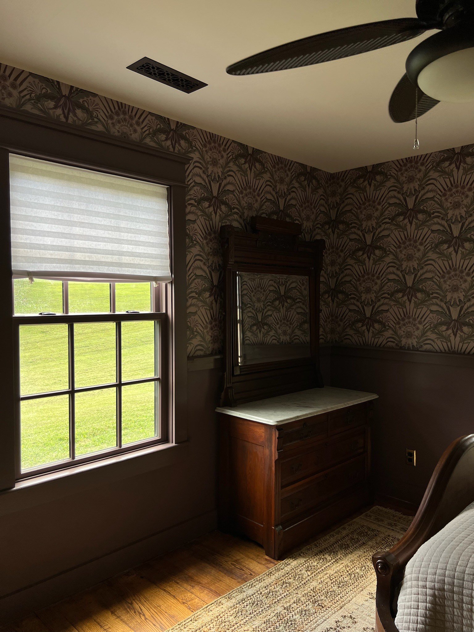 Interior view of a room with a window showing a green lawn outside, wallpaper with floral pattern, a wooden dresser with a mirror, a ceiling fan, and a beige rug.