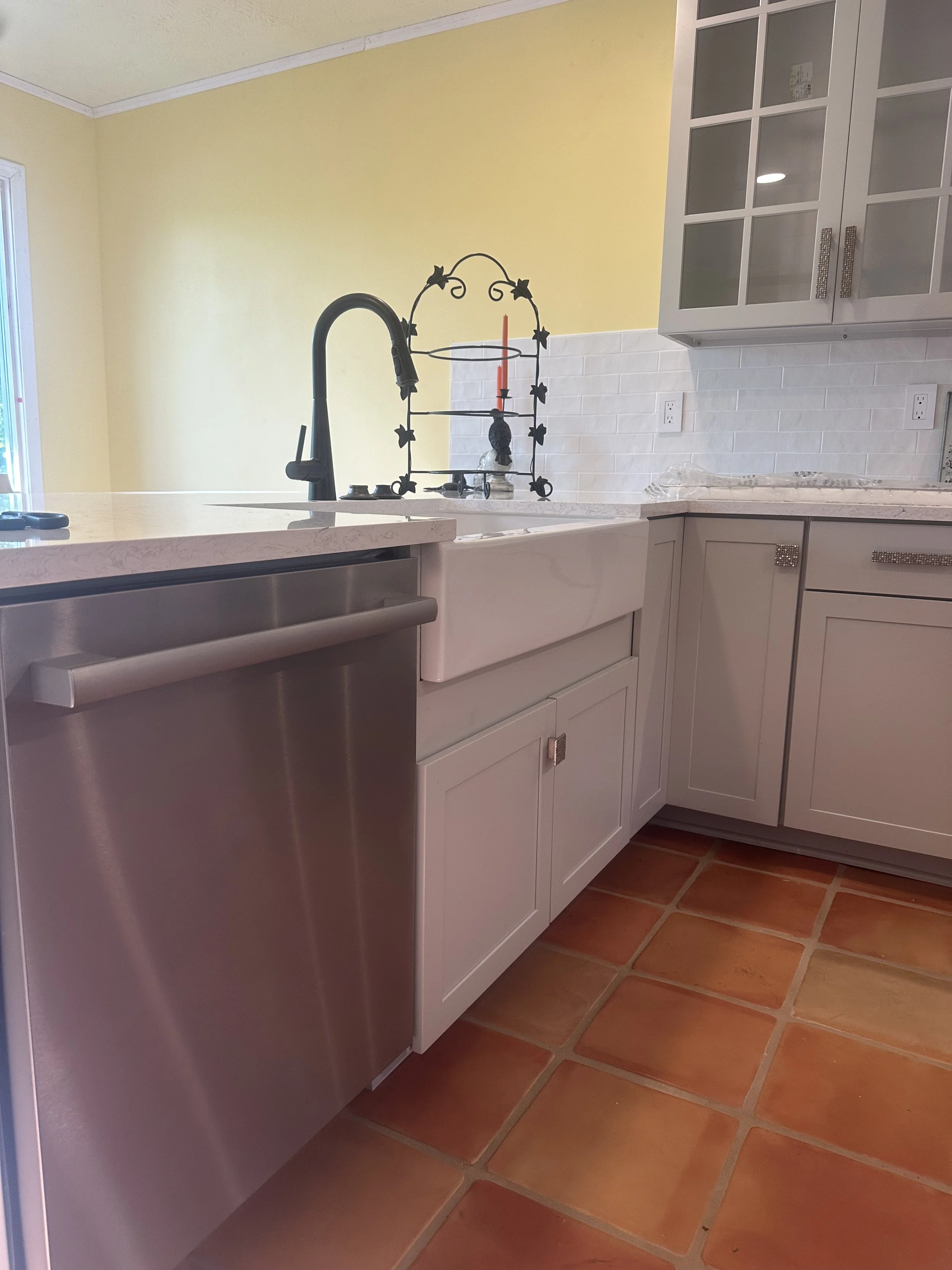 Kitchen with yellow wall, white cabinets, stainless steel dishwasher, black faucet, white farmhouse sink, and terracotta tile floor.