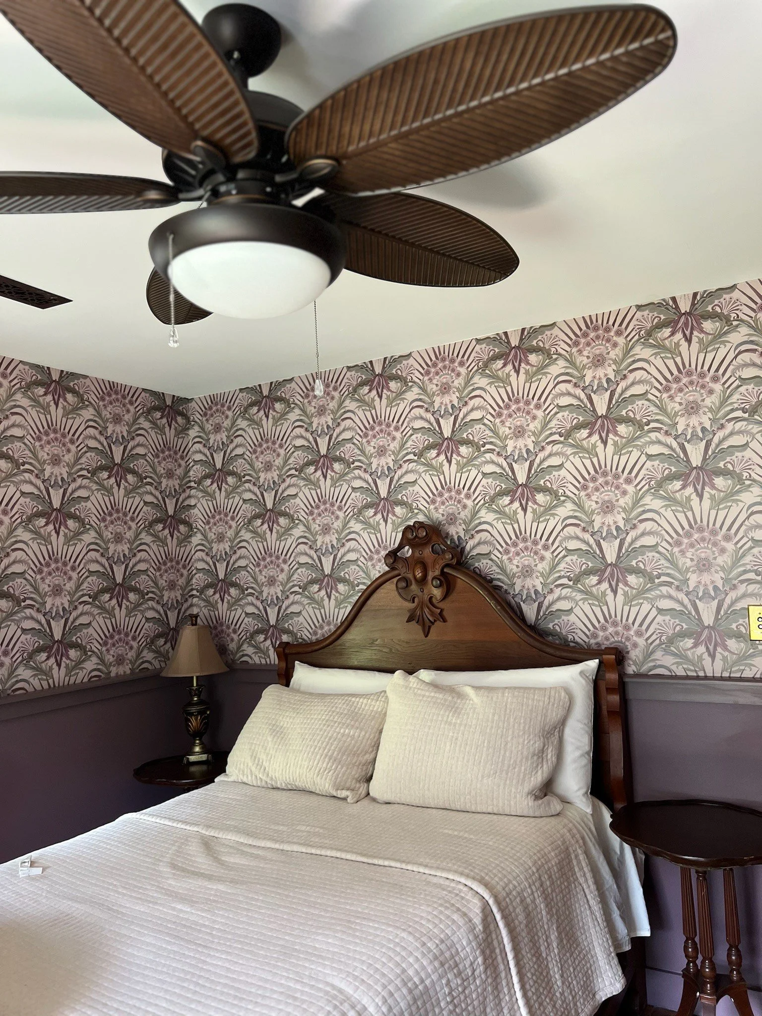 A bedroom with a floral wallpaper, a wooden bed with decorative headboard, two pillows, a side table with a lamp, and a ceiling fan with wooden blades and a light fixture.