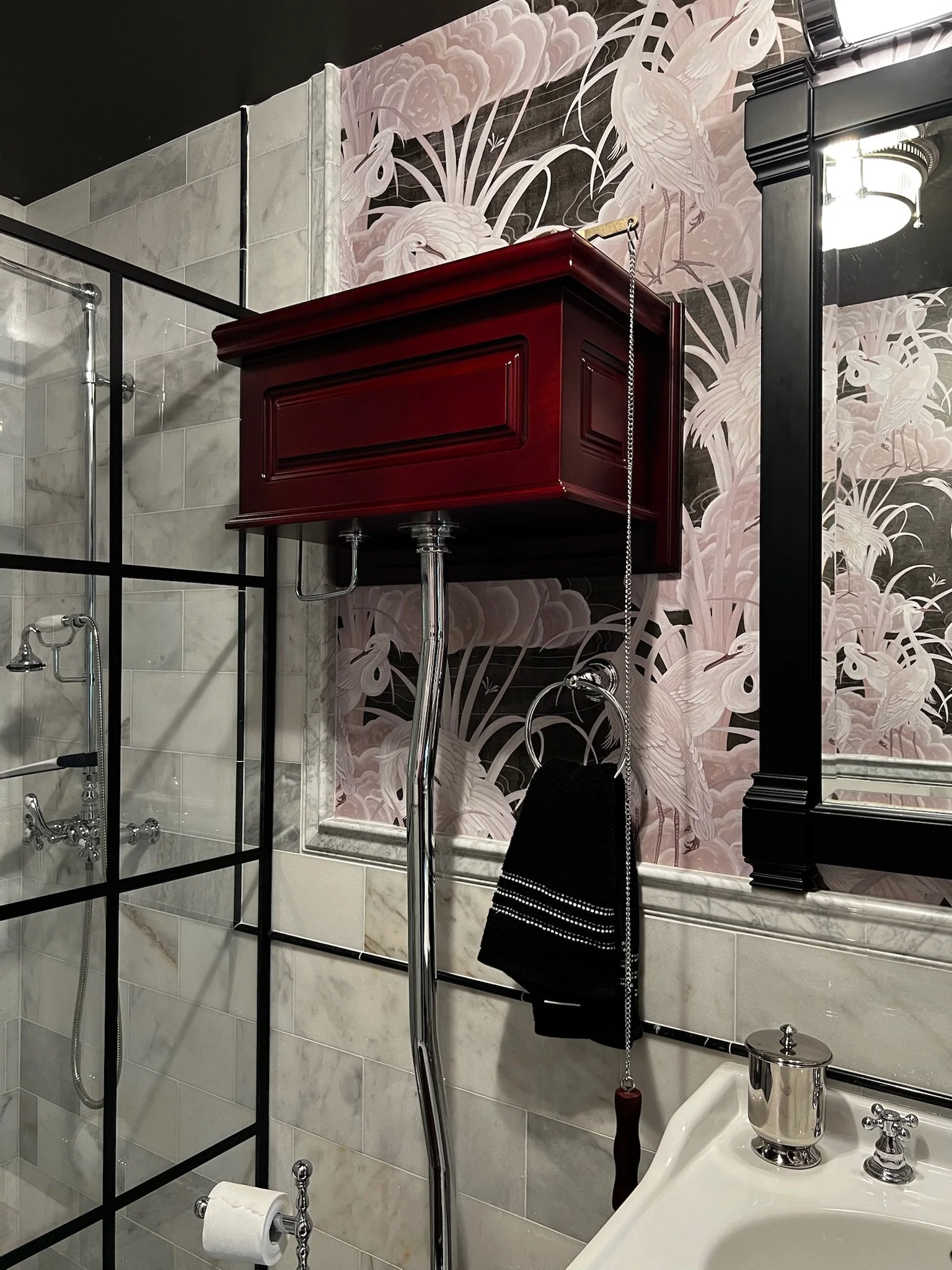 A bathroom with a red toilet tank mounted on the wall, a black and white towel hanging on a ring, a mirror with a black frame, a marble sink with a silver soap dispenser, and floral wallpaper featuring storks and pink flowers.