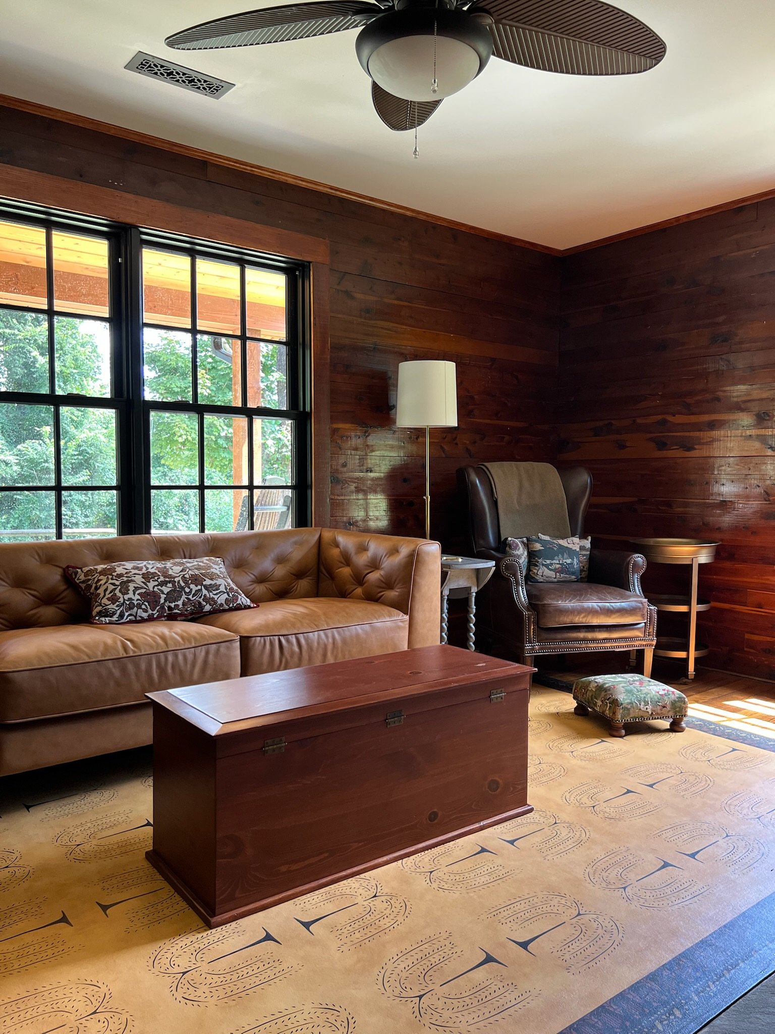 Living room with wooden walls, a large window, a leather couch, an armchair, a wooden chest, and a patterned rug, with a ceiling fan and floor lamp.