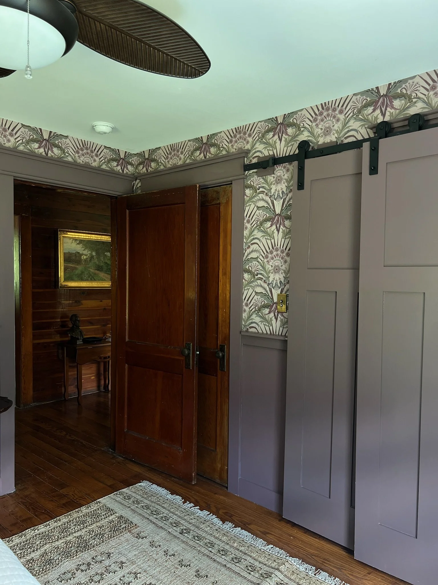 A room with a vaulted ceiling, wallpaper with floral pattern, a ceiling fan, grey sliding barn door, and a wooden door. Part of a rug and hardwood floor are visible.
