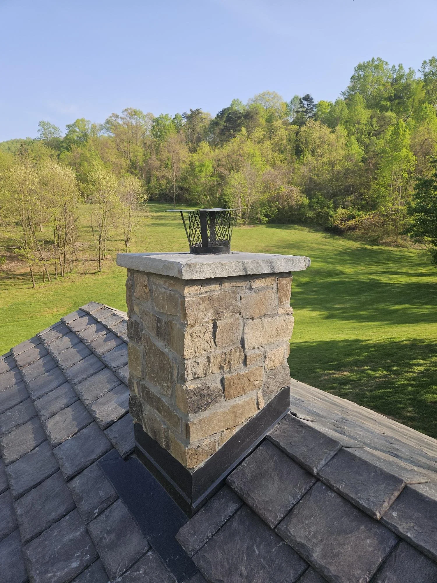 A stone chimney on a shingled roof with a metal chimney cap, in a grassy field with trees and a blue sky in the background.