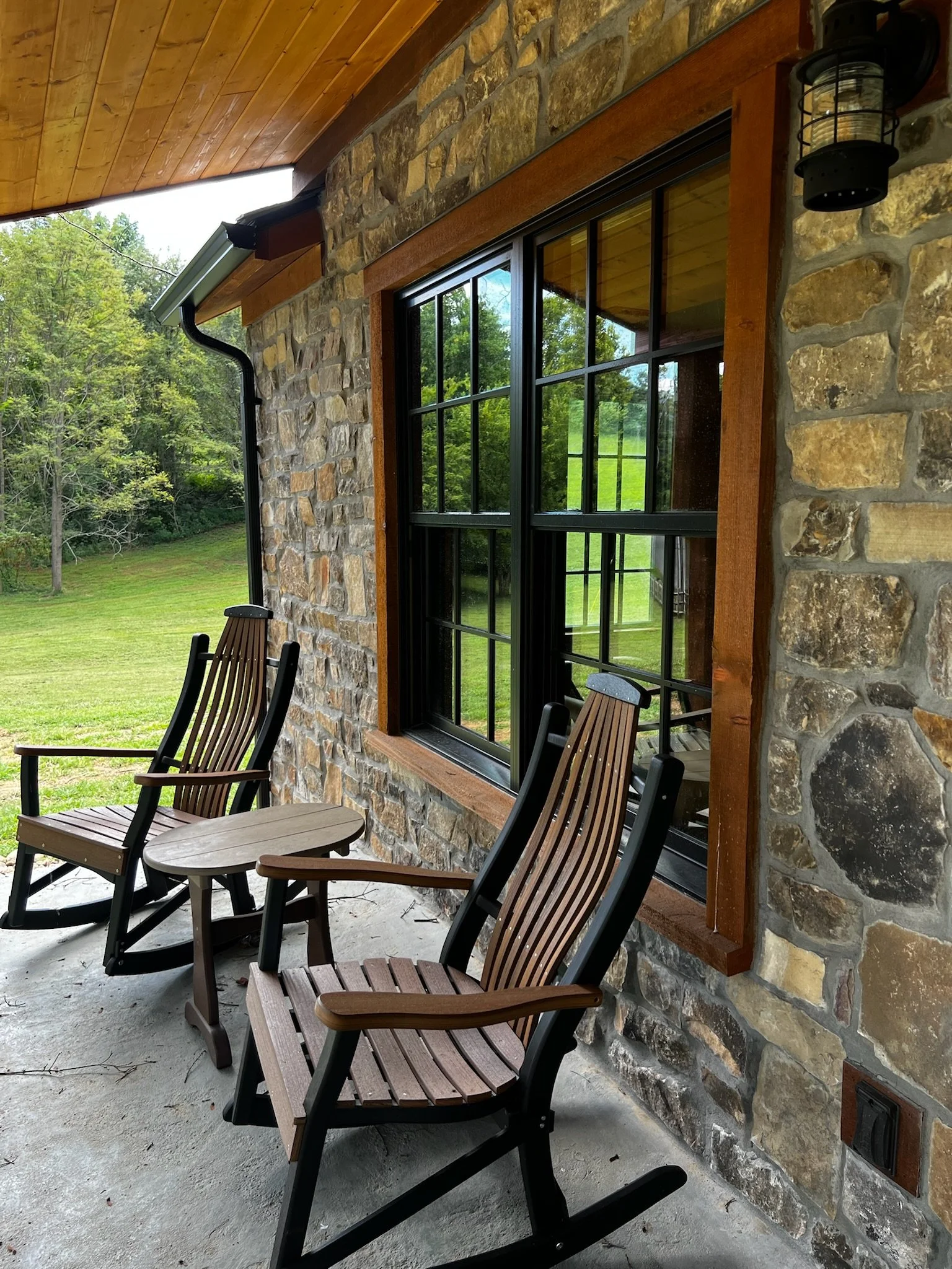 Front porch with two wooden rocking chairs and a small side table against a stone wall and large window, surrounded by green grass and trees.