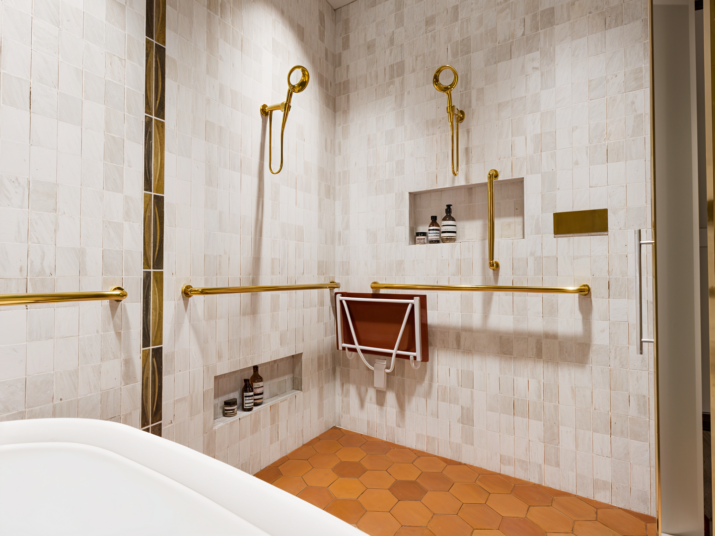 Accessible walk-in shower with beige tiled walls, gold grab bars, two shower heads, and shampoo bottles in recessed shelf.