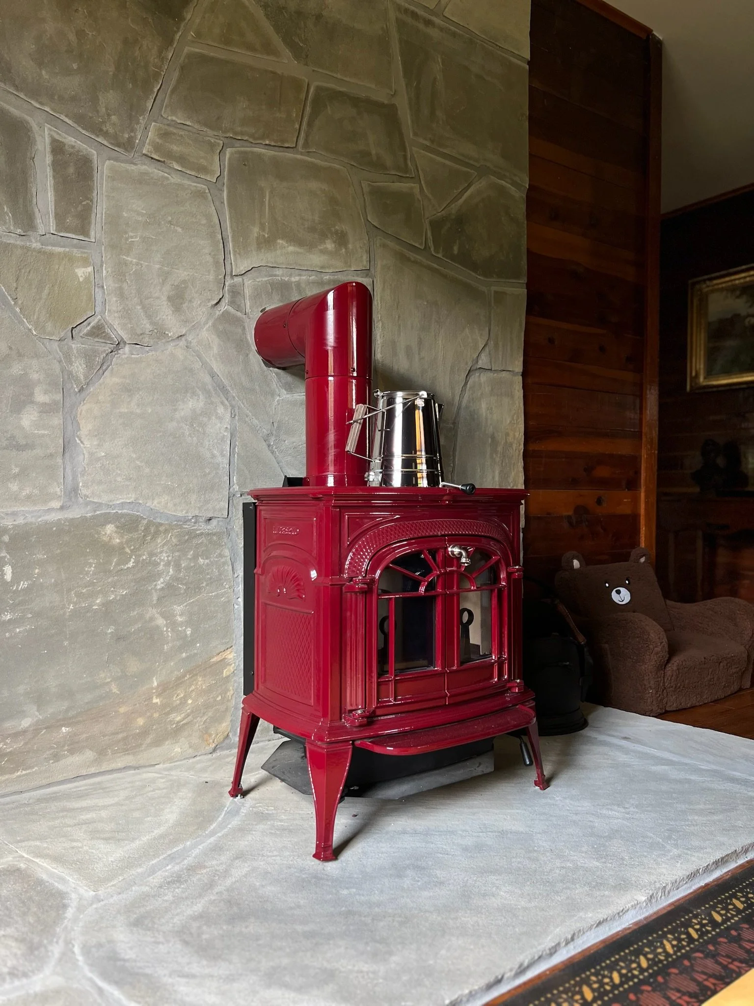 Red vintage wood stove on a stone hearth in a cozy living room with brown wood walls and a bear-shaped cushion on a small brown sofa.