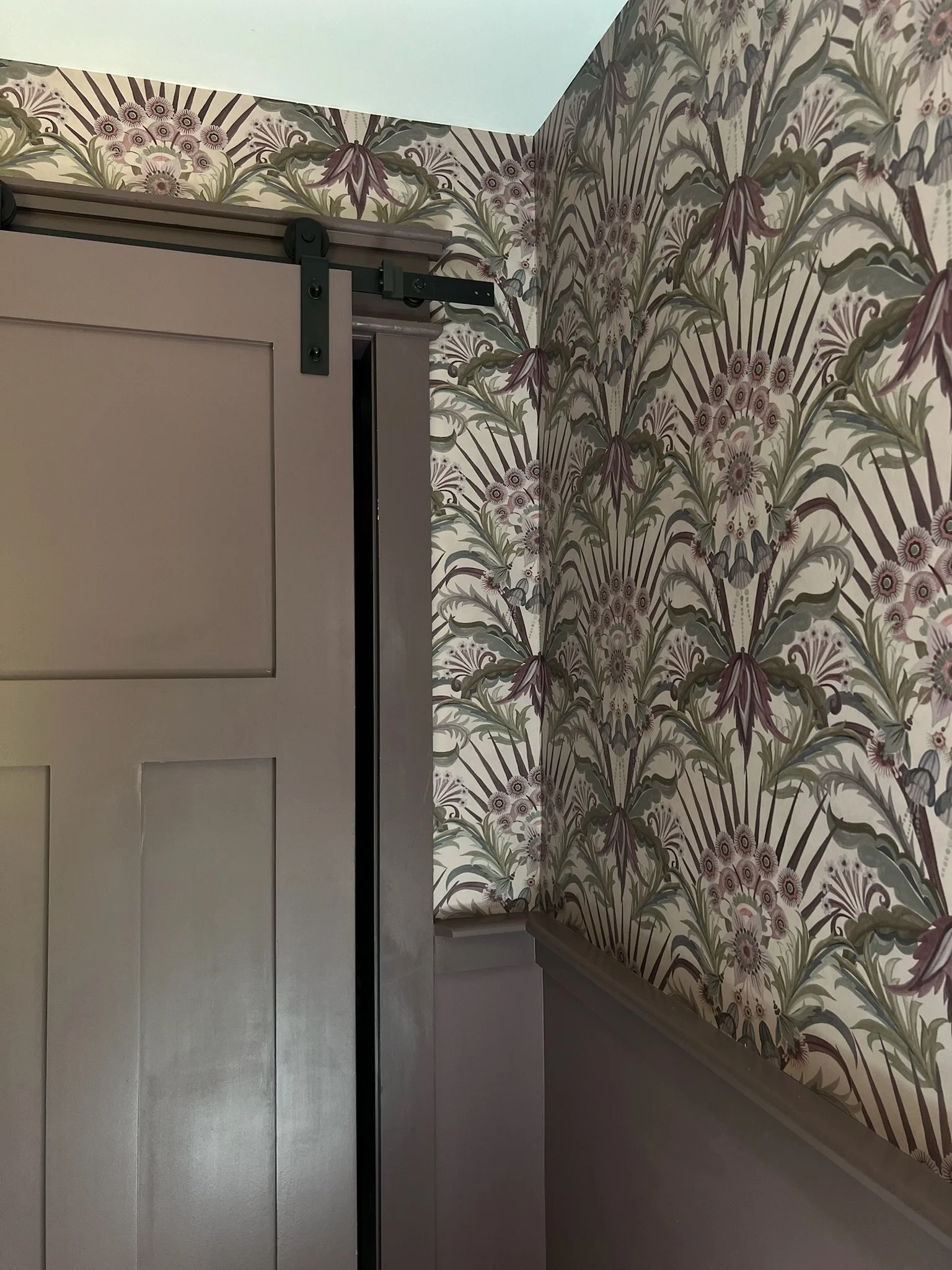 Close-up of a sliding barn door with dark black hardware, installed in a room with floral patterned wallpaper in shades of beige, brown, and muted purple.
