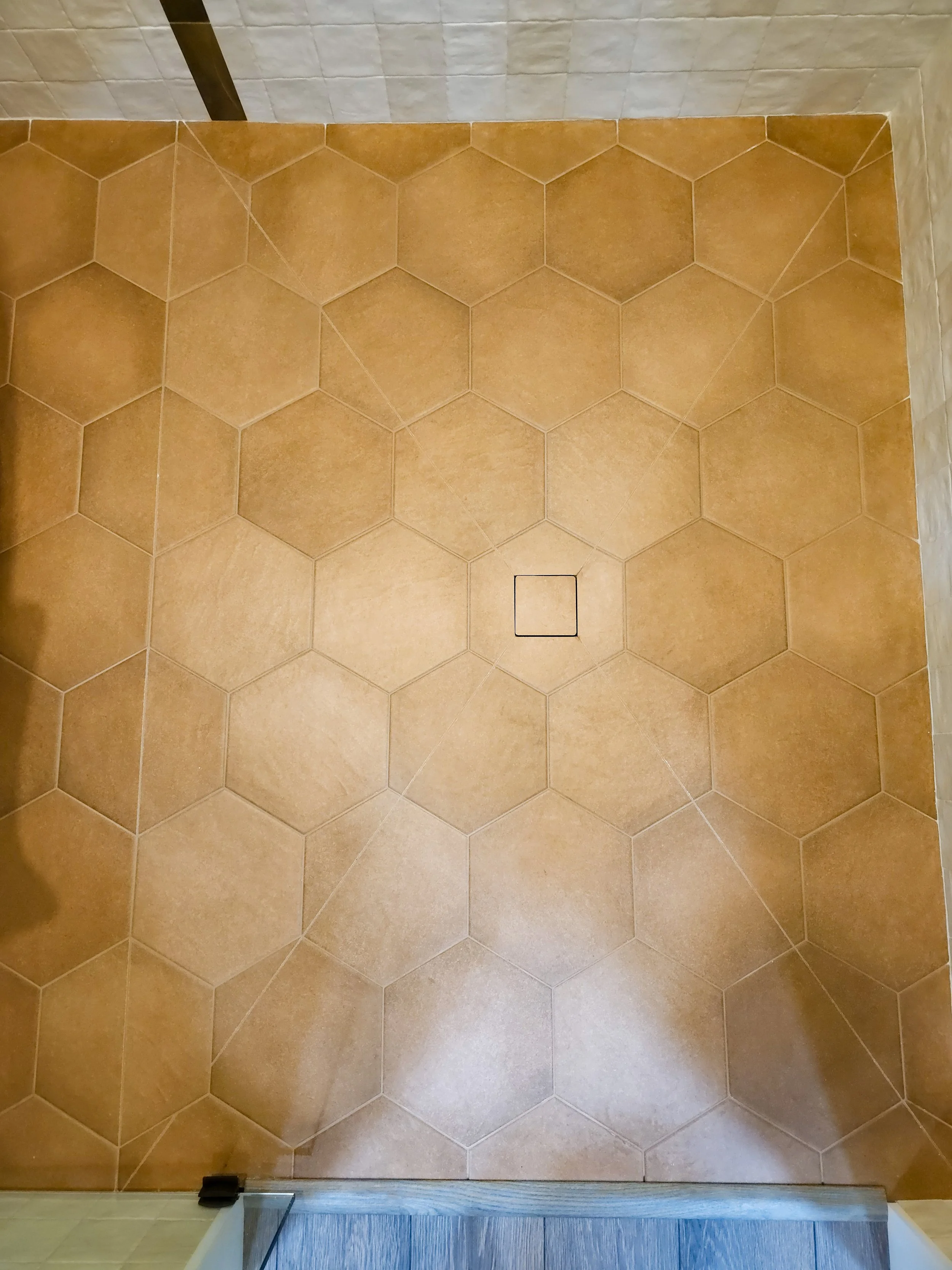 A top-down view of a tiled bathroom or shower floor with large hexagonal orange-brown tiles, a square drain in the center, and a partial wooden threshold at the bottom.