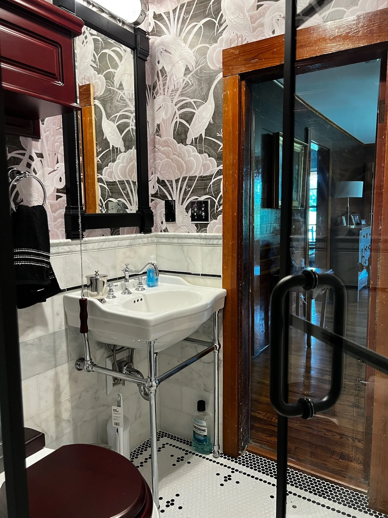 Bathroom with floral wallpaper, a white sink, a burgundy toilet, and a glass door leading to a room with dark wood paneling and a blue ceiling.