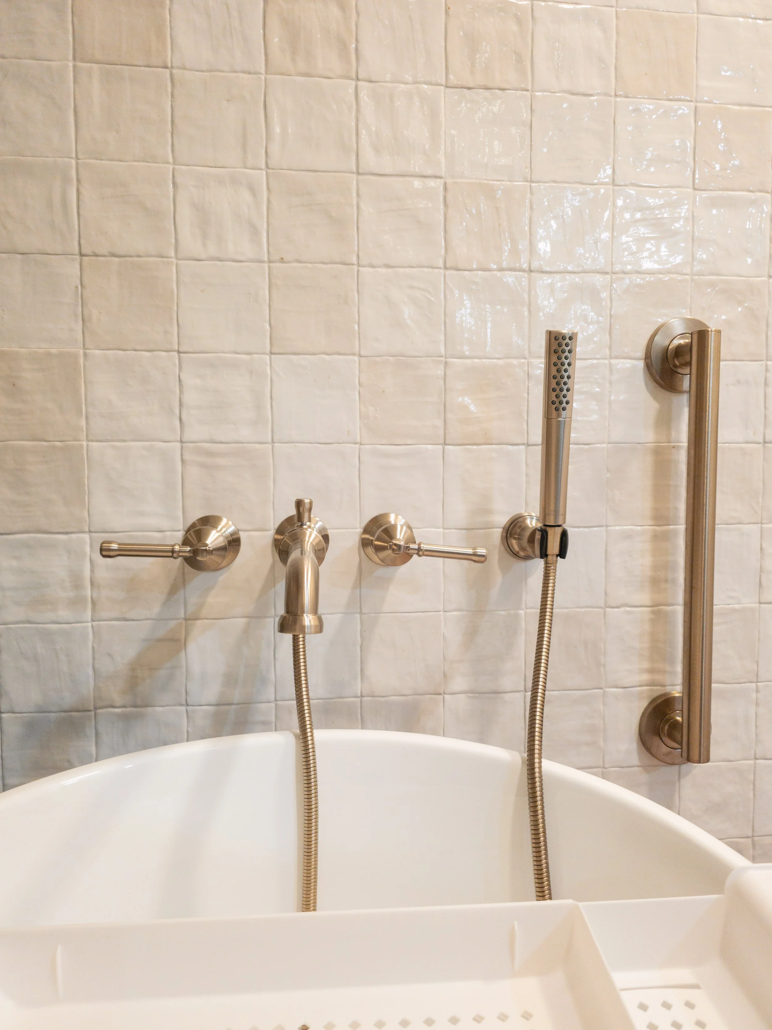 Bathroom with beige tiled wall, bathtub with stainless steel fixtures, an adjustable handheld shower head, and grab bars.
