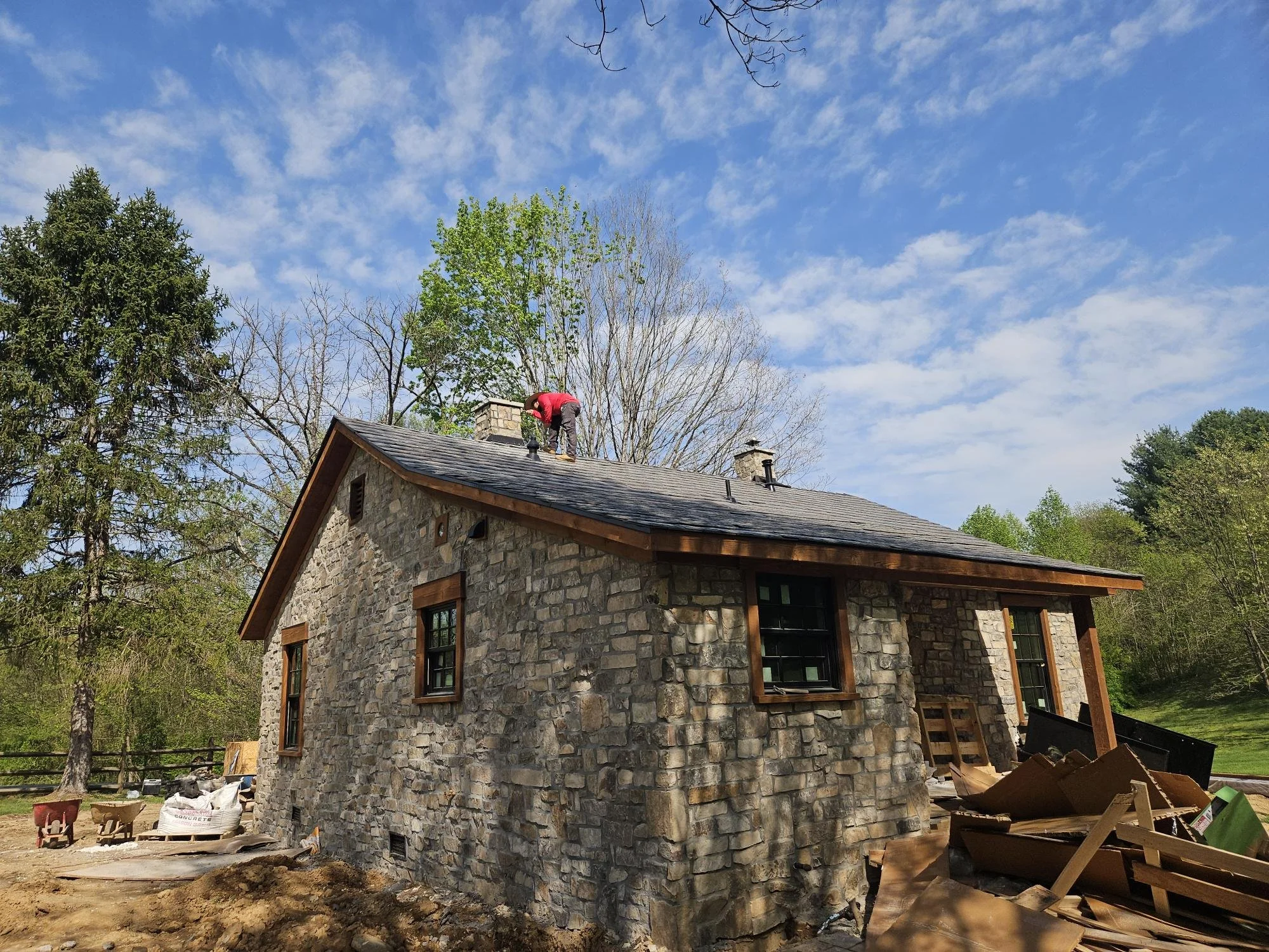 A person in a red shirt working on the roof of a stone house during construction or renovation, with piles of lumber and construction materials outside, surrounded by trees and under a partly cloudy sky.