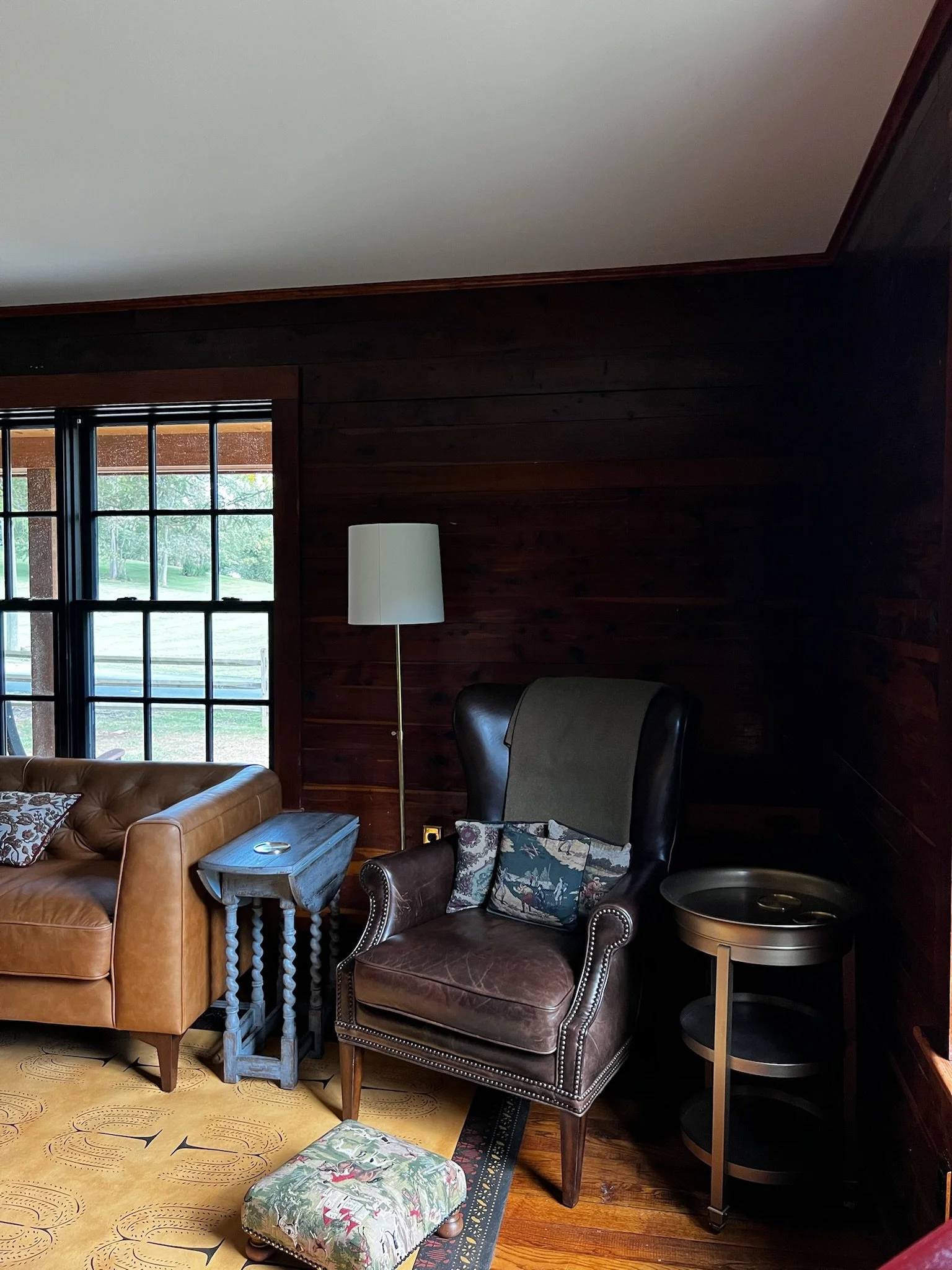 Living room with dark wooden walls, a large window, a leather armchair with a decorative pillow, a tan leather sofa, a small side table, a tall floor lamp, a small round side table, and a patterned footstool.