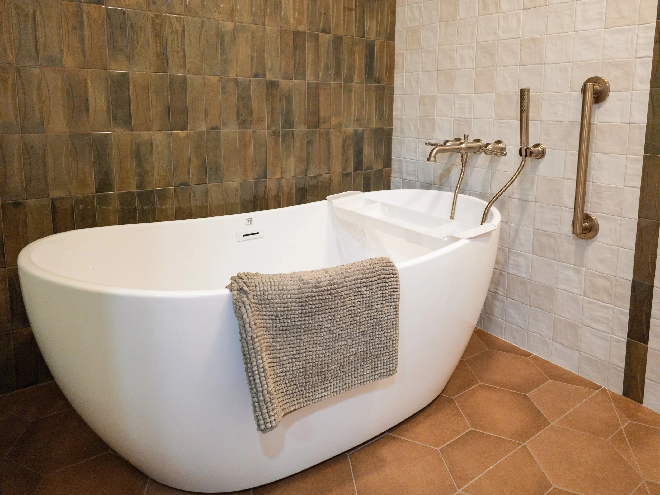 A modern bathtub with a gray towel hanging over the side, set against a wall with brown and white tiled walls and bronze grab bars.